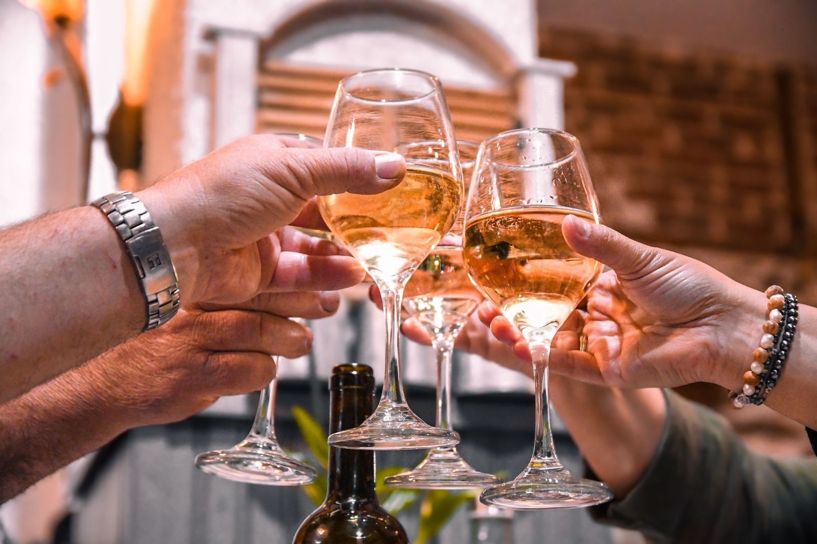 Two pairs of hands clinking wine glasses together over a table with a wine bottle, at an outdoor evening gathering