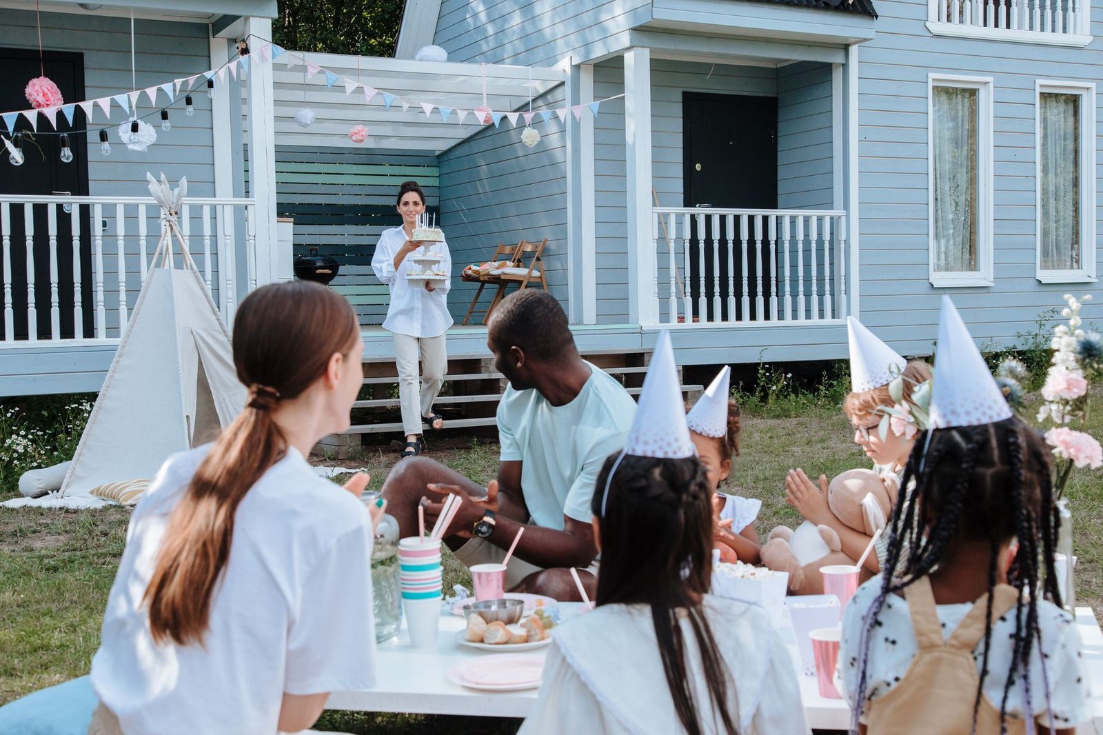 A woman carrying a two-tier birthday cake with lit candles toward a group of adults and children in party hats seated at an outdoor table decorated with bunting