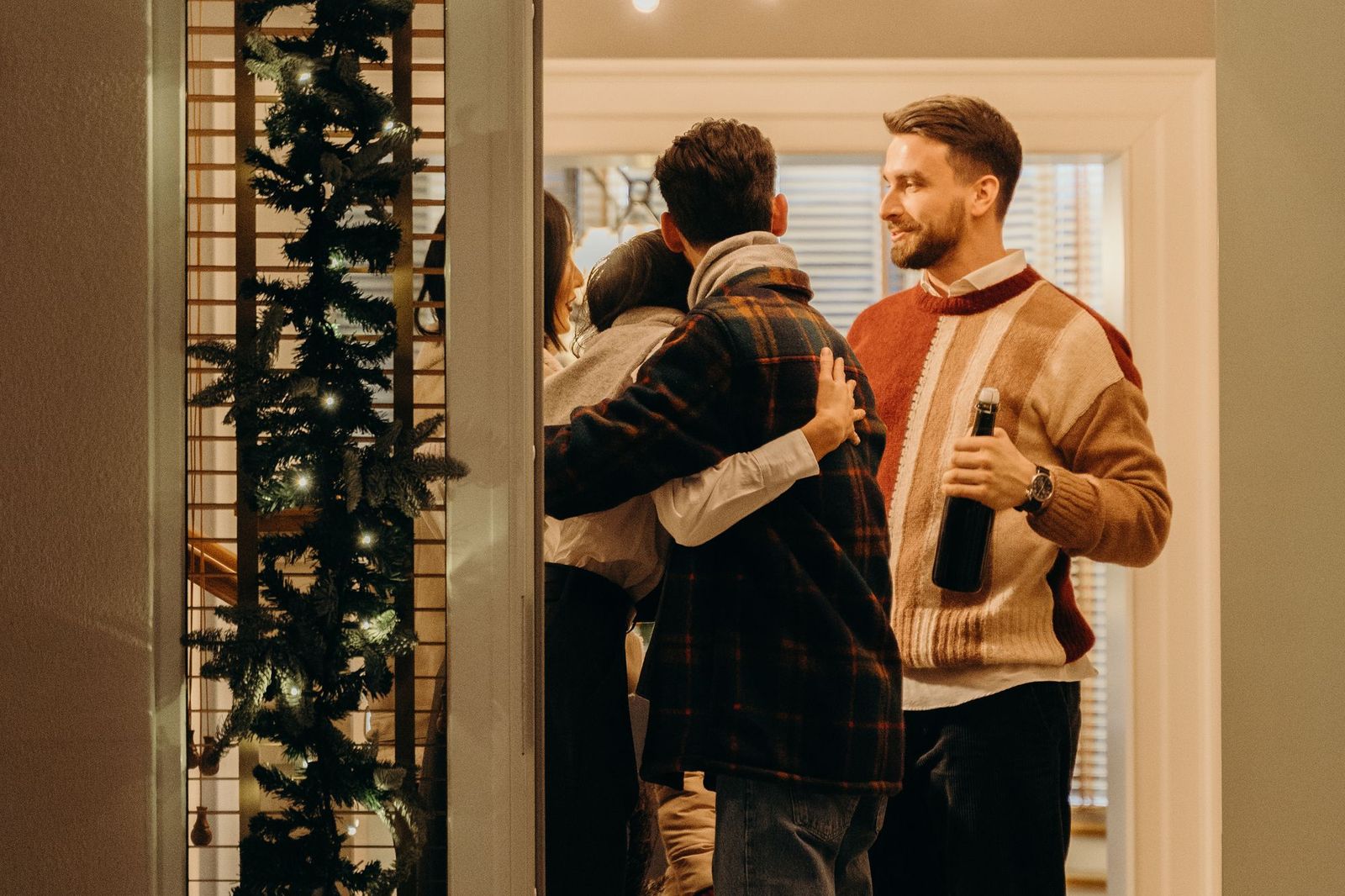 A small group hugging warmly in a doorway as a new guest arrives, one man standing slightly apart holding a bottle of wine