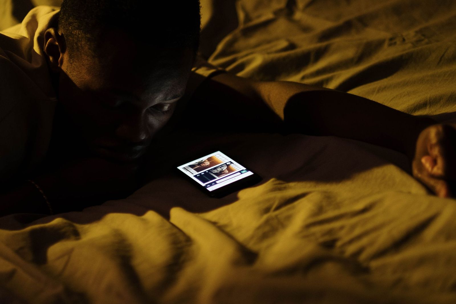 A man lying in bed at night, his face lit only by the glow of a phone showing a busy chat conversation, with another person visible sleeping beside him