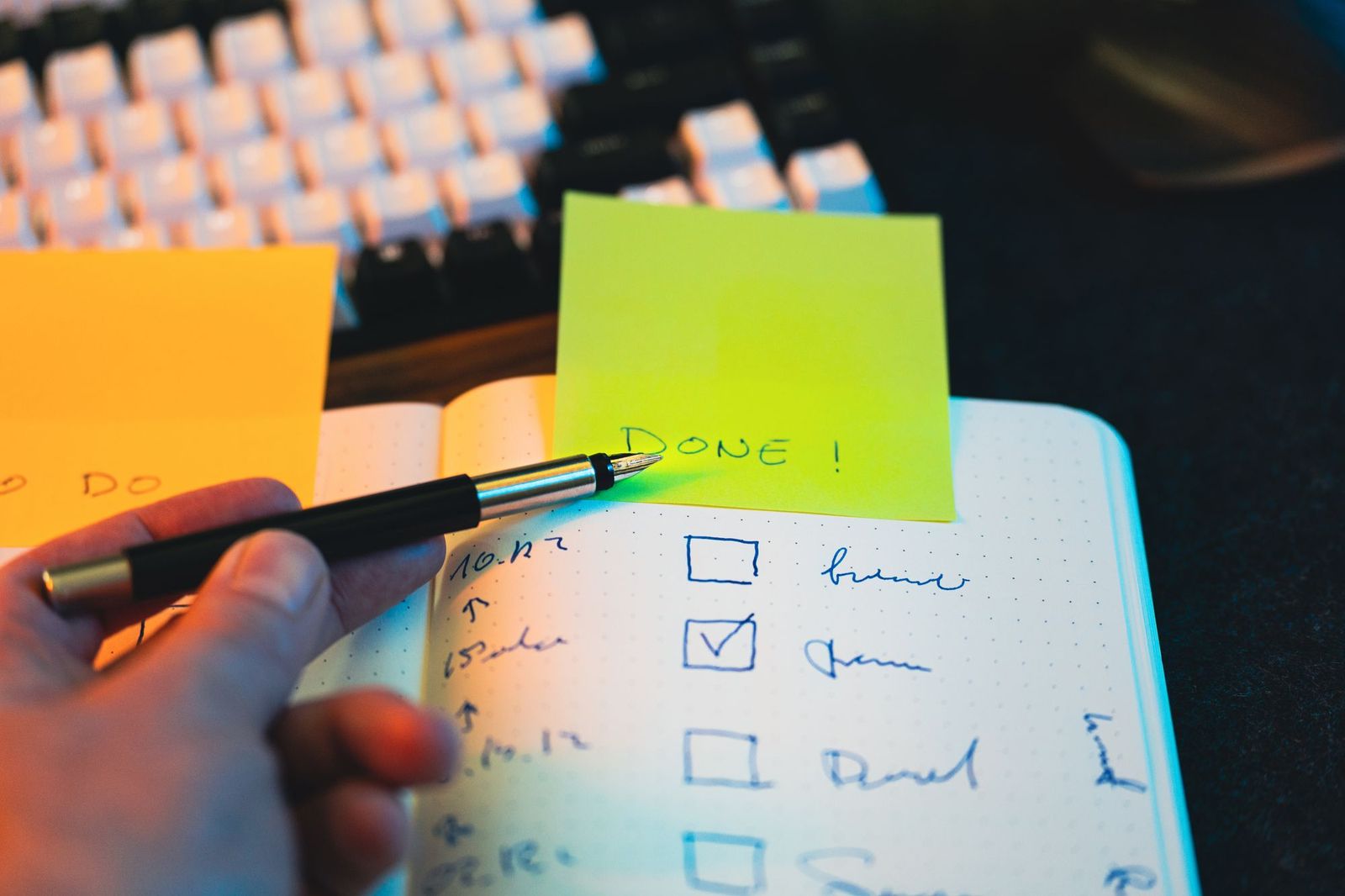 A hand holding a pen over an open notebook with a checklist, a green sticky note reading DONE stuck beside it