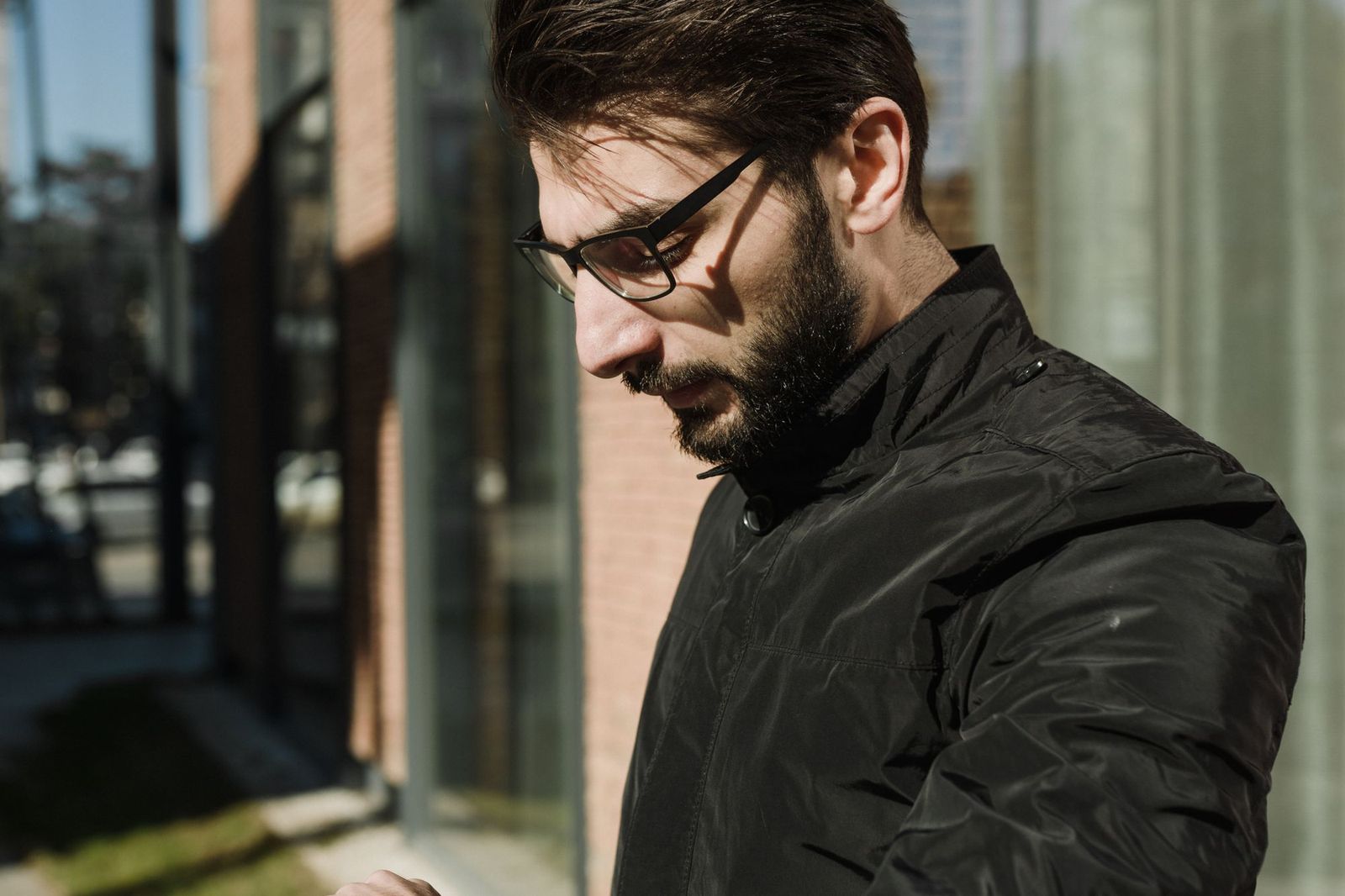 A man with glasses and a beard standing outside a glass storefront, looking down thoughtfully with sunlight catching his face