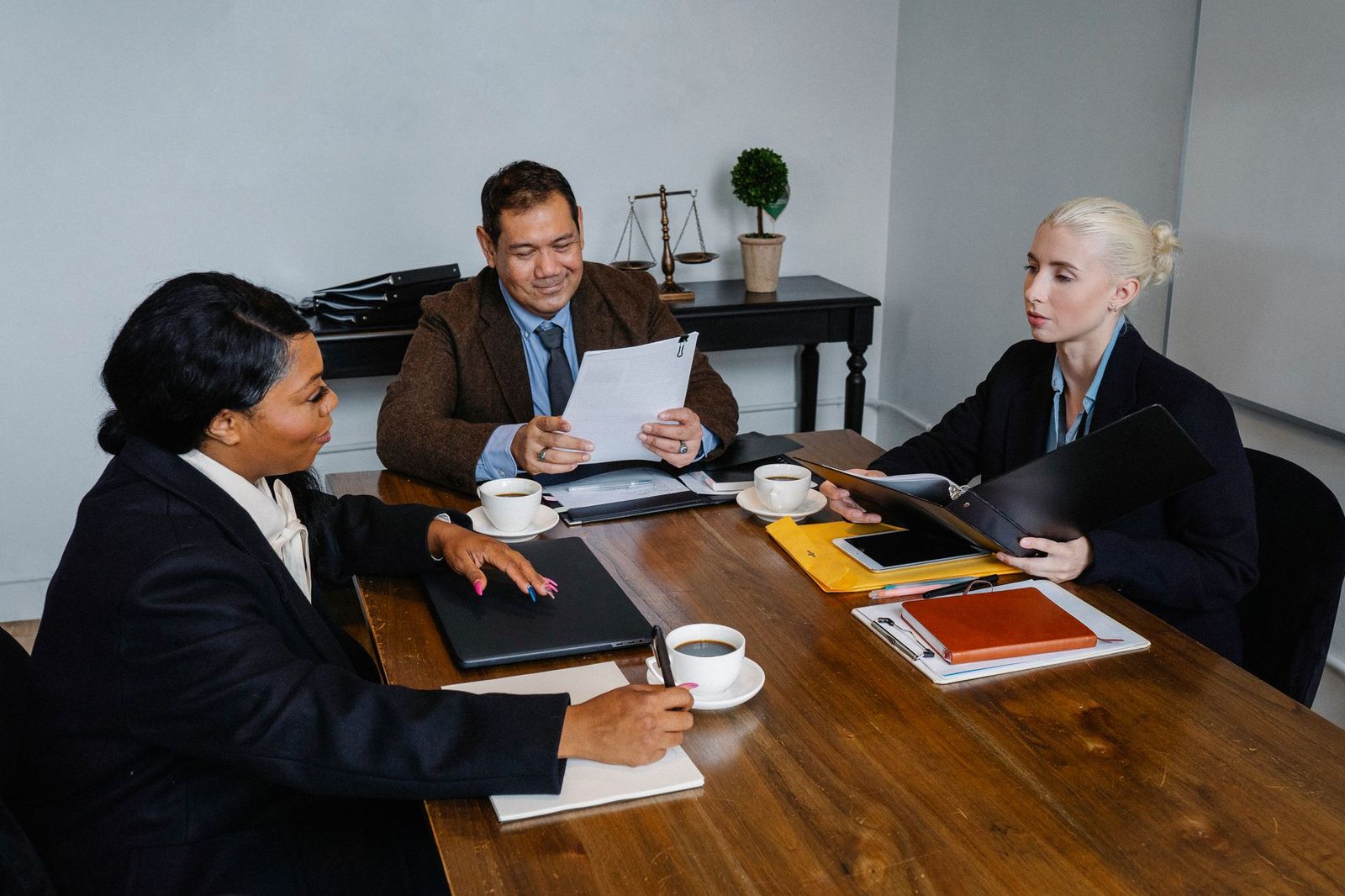 Three people in formal business dress sitting around a wooden meeting table with coffee cups, one reading a printed page while another holds an open black folder