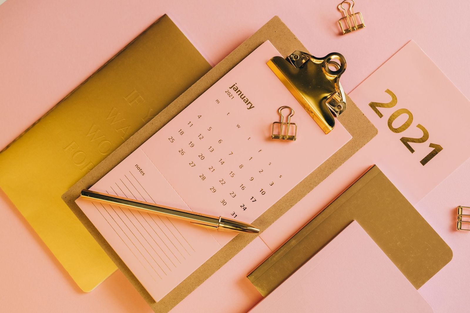 A flat lay on a pink background of a clipboard holding a January desk calendar page with a notes section, a gold pen laid across it and gold paper clips beside