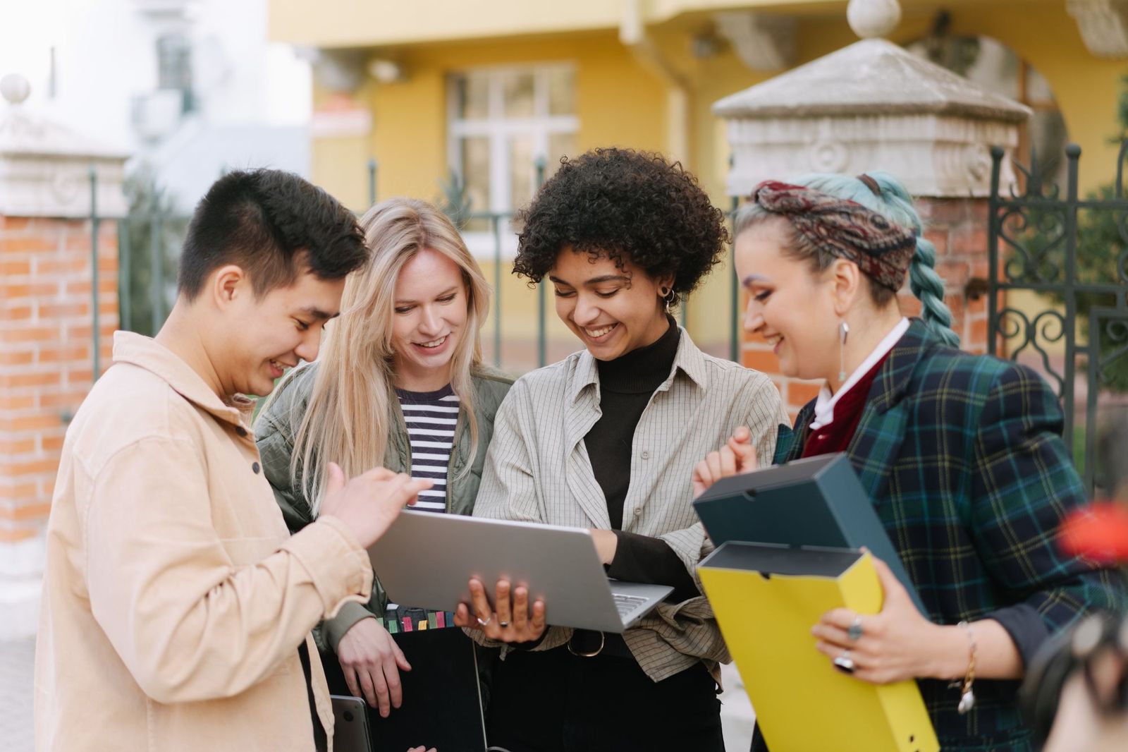 Four students standing outside a building, leaning in together to look at a laptop one of them is holding open, folders under their arms