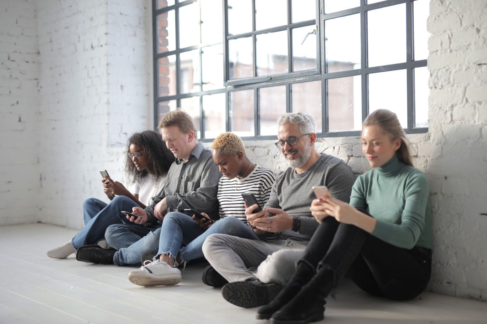 Five people sitting in a row on the floor against a white brick wall by a tall window, each looking down at their own phone