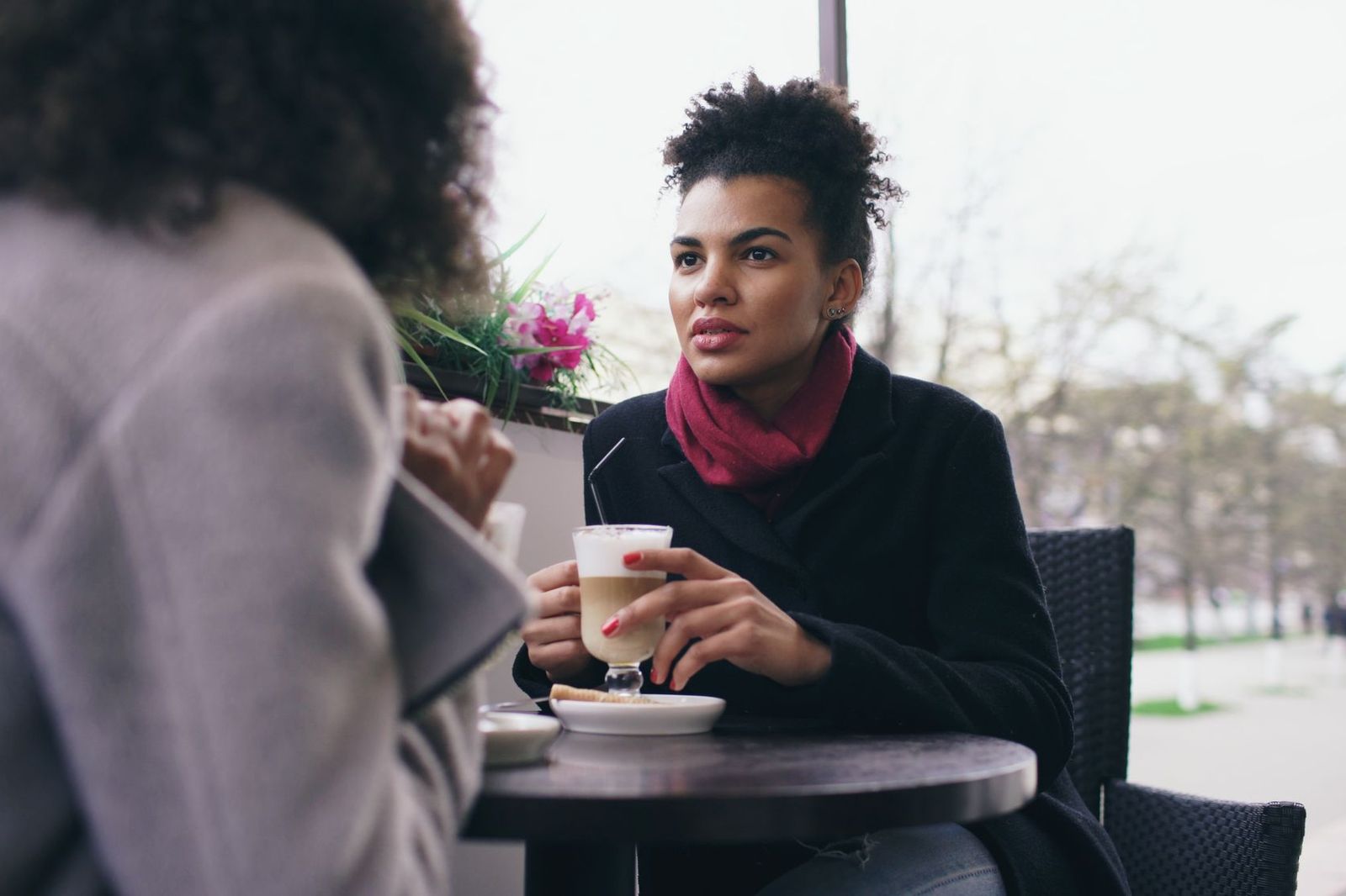 Two women sitting at an outdoor cafe table with coffee, one listening closely as the other talks, a window box of pink flowers behind them