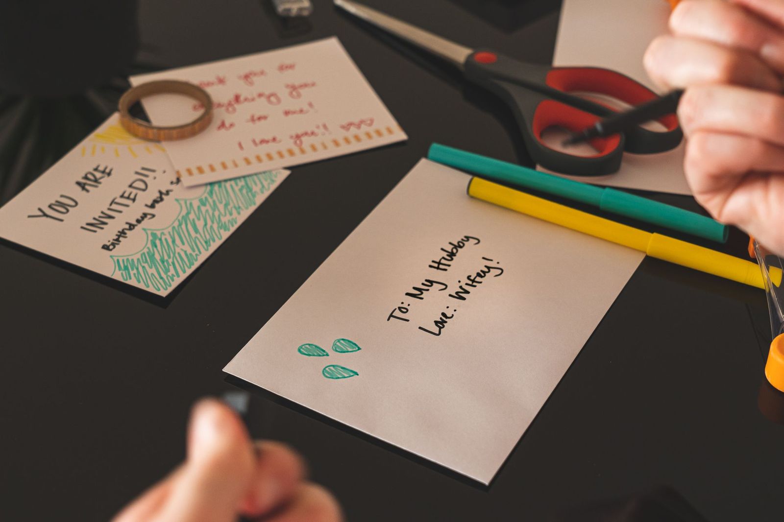 A handmade birthday invitation card reading YOU ARE INVITED lying on a dark desk beside felt tip pens, scissors and a sealed envelope addressed in marker pen