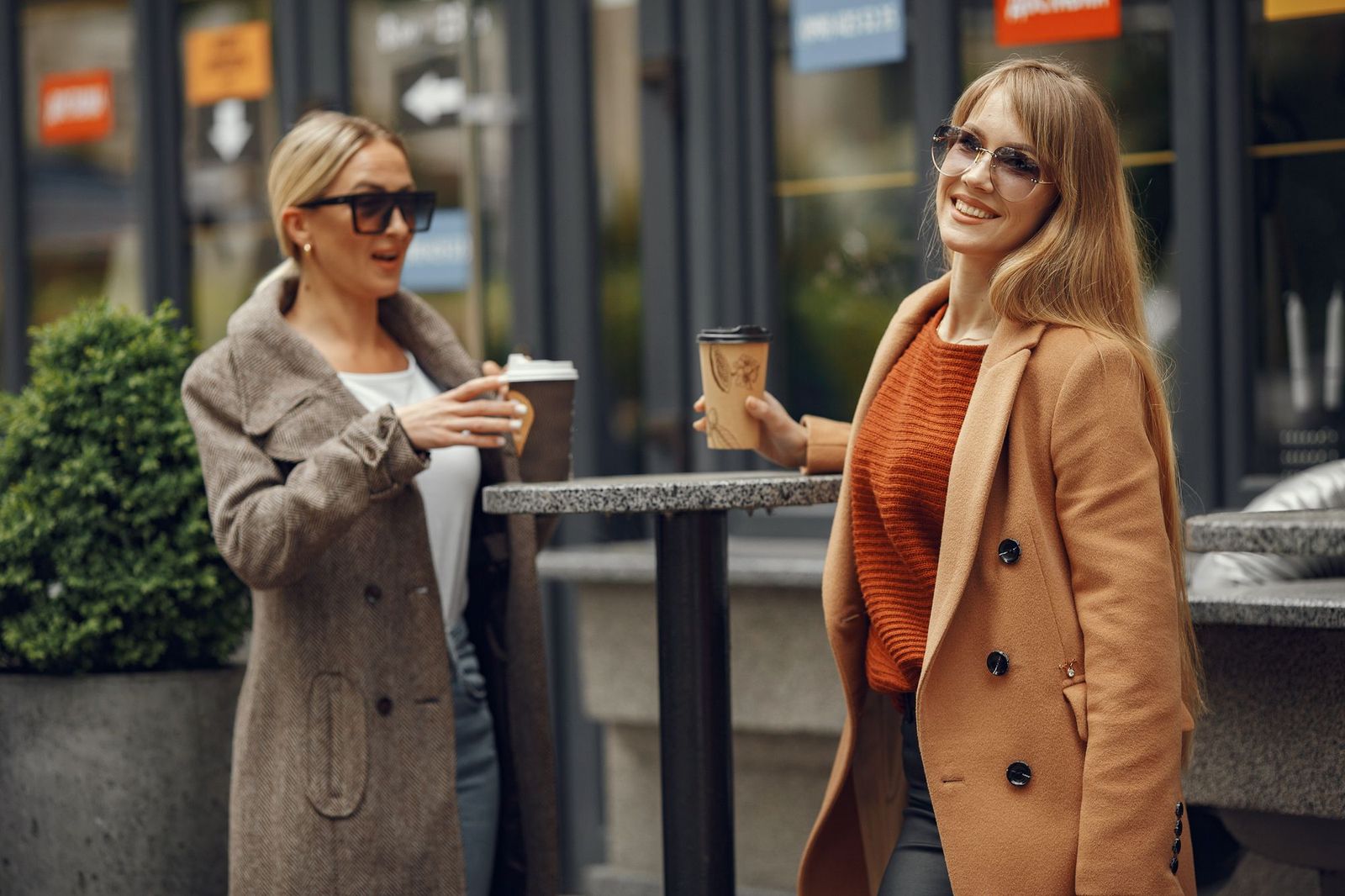 Two women standing at an outdoor cafe table on a city street, both holding takeaway coffee cups, one talking and gesturing while the other looks toward the camera