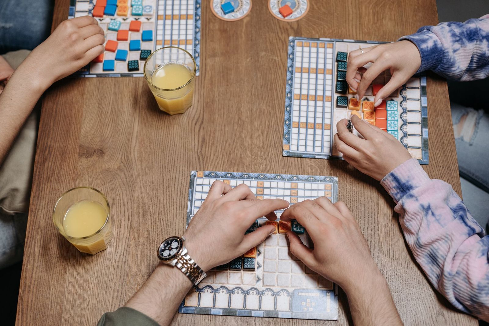 Overhead view of three people playing a tile-based board game around a wooden table, with two glasses of juice beside the boards