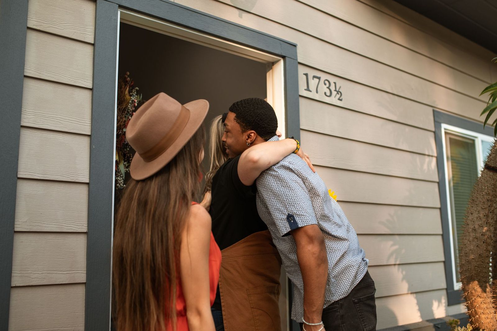 Two people embracing in a doorway of a house while a third guest in a wide-brimmed hat waits behind them