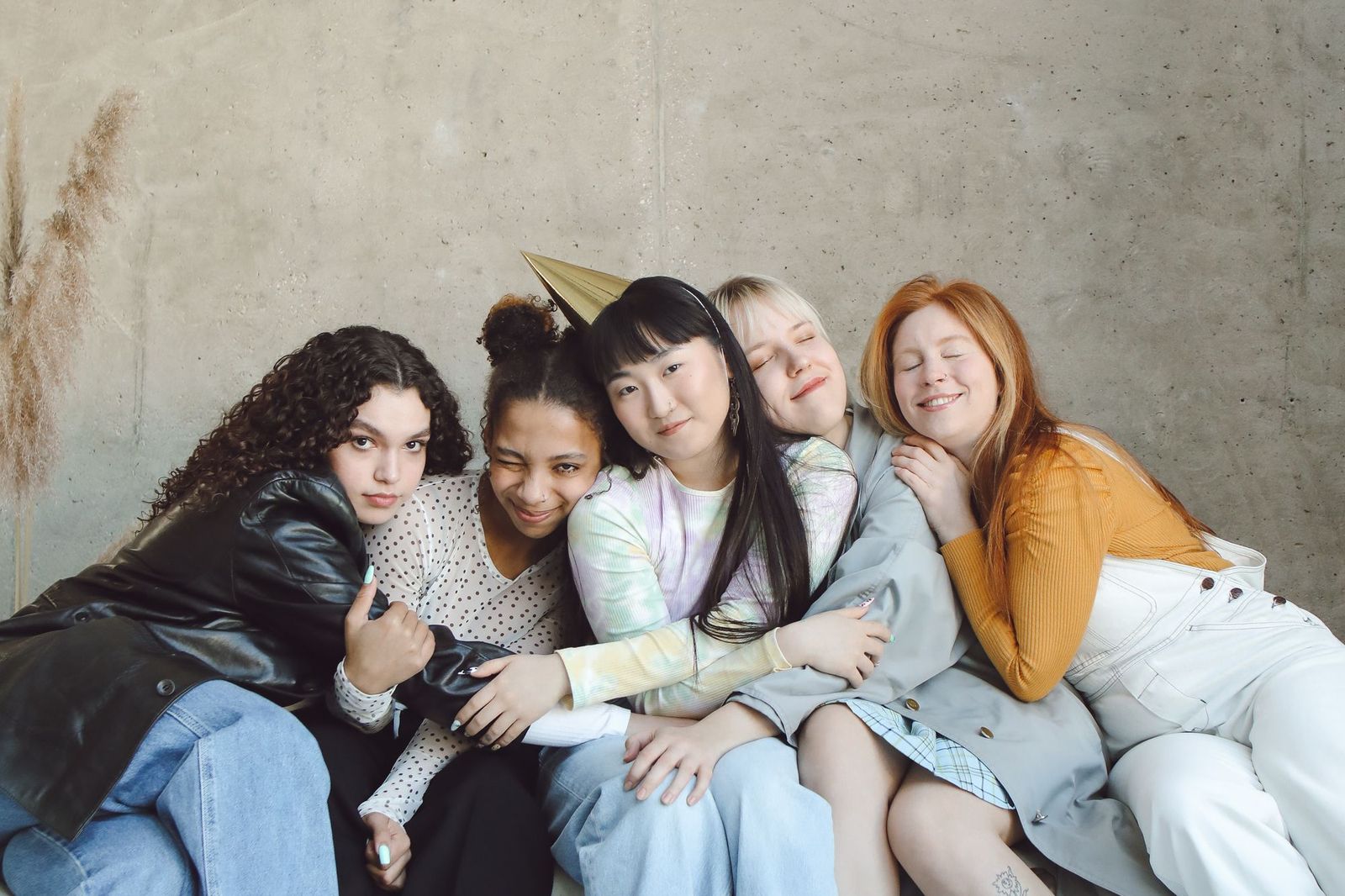 Five friends sitting squashed together against a concrete wall with their arms around each other, one wearing a gold party hat