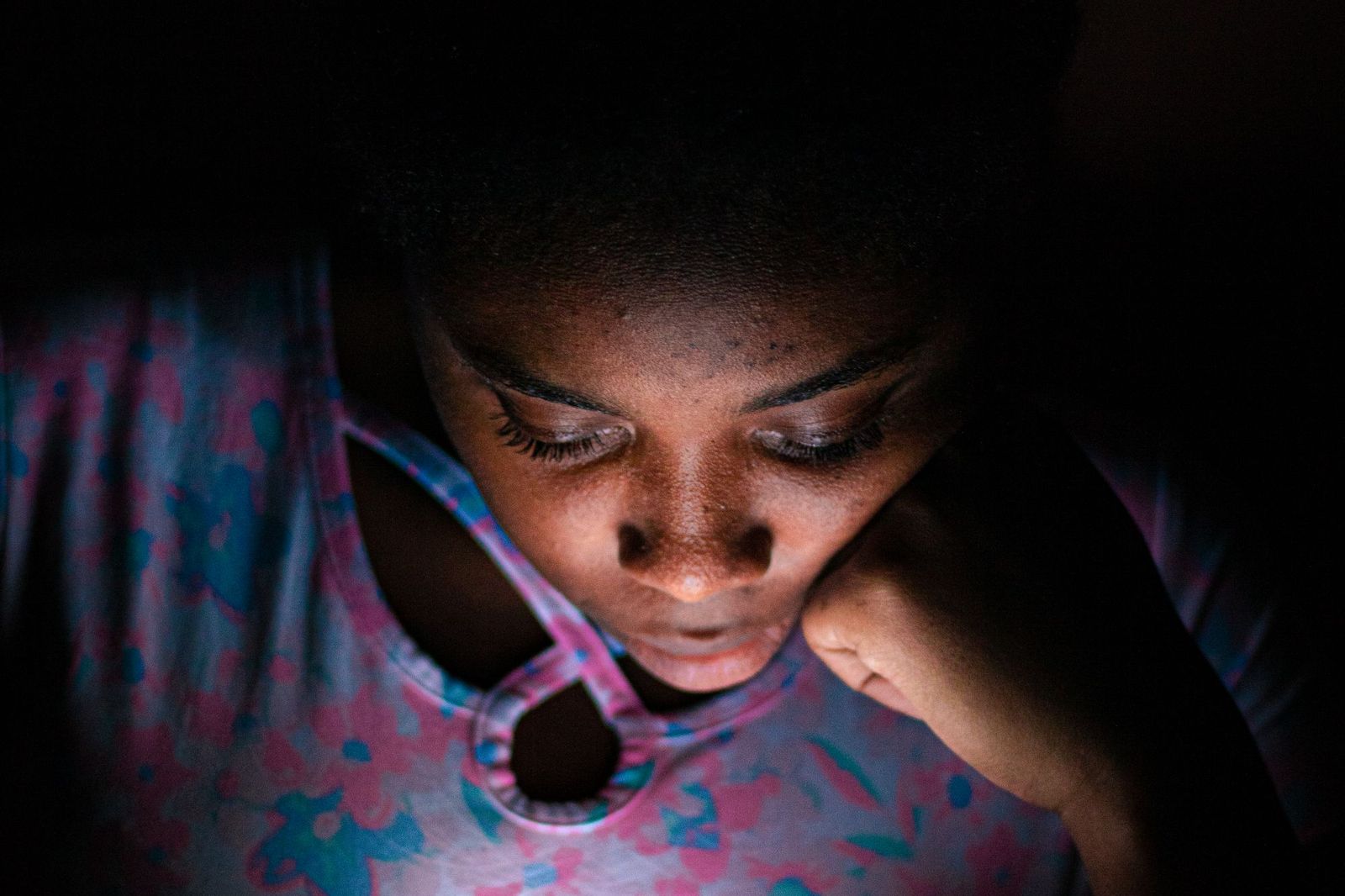 Close-up of a young woman alone in a dark room, her face lit from below by the glow of a phone screen