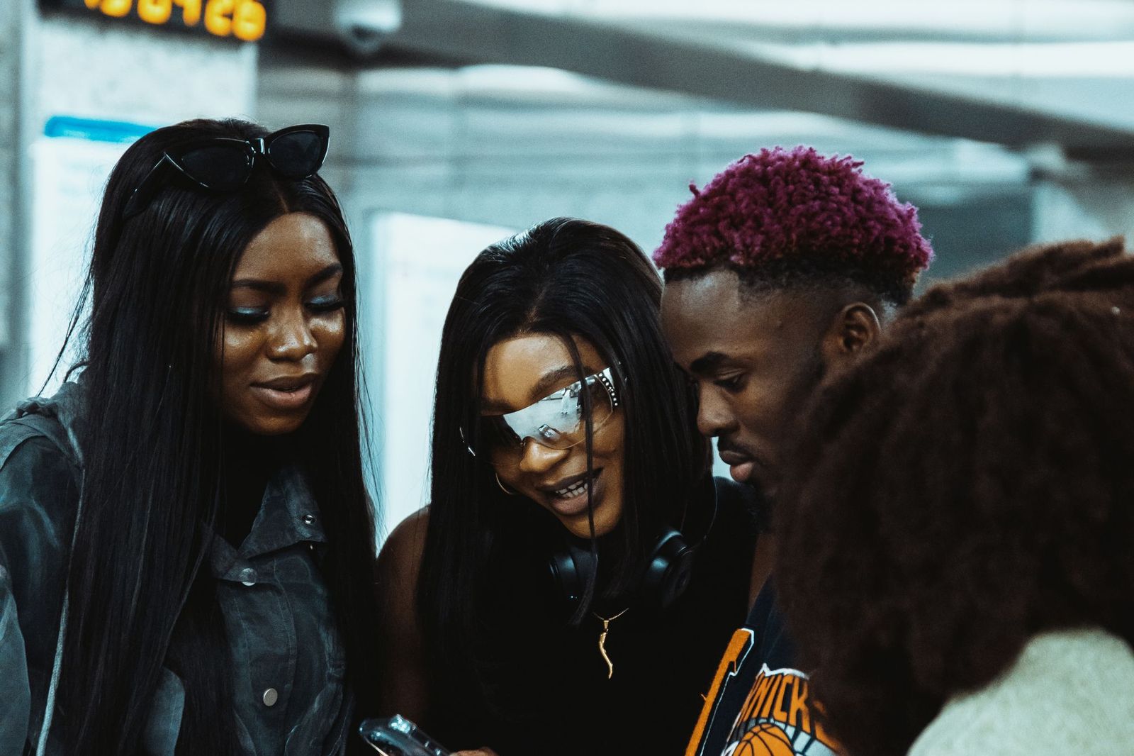 Three friends standing close together outdoors in a station concourse, looking down at one phone held by the person in the middle