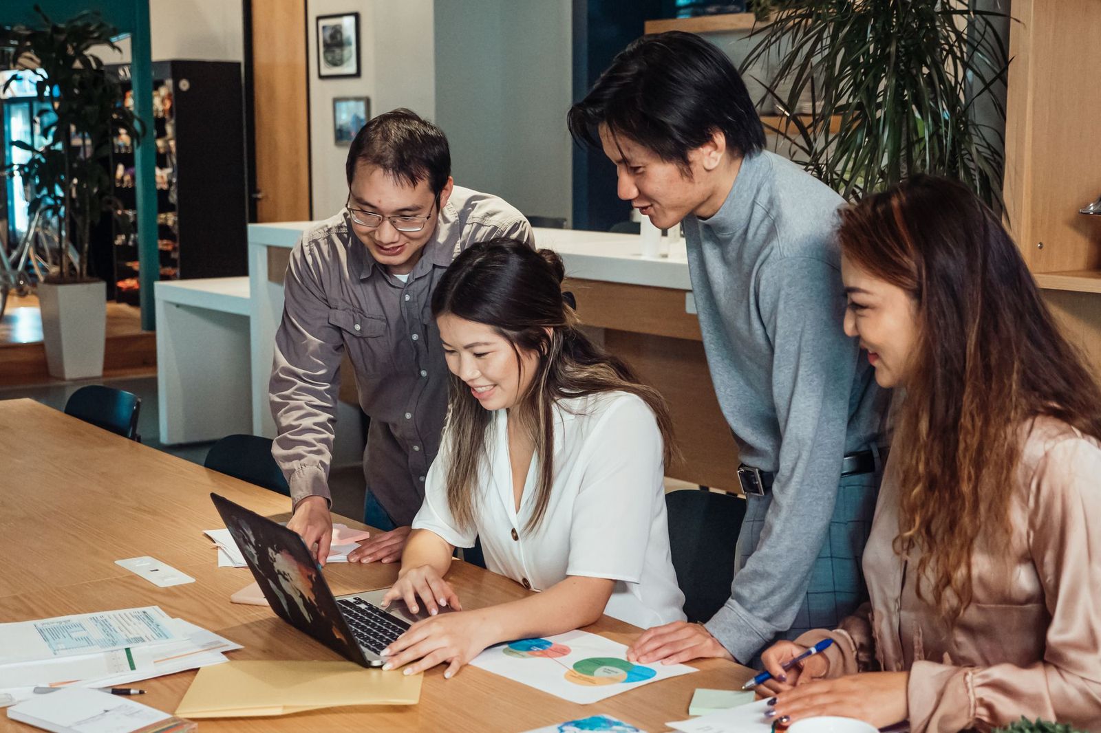 Four colleagues gathered around a laptop at a wooden desk, one typing while the others lean in to look at the screen