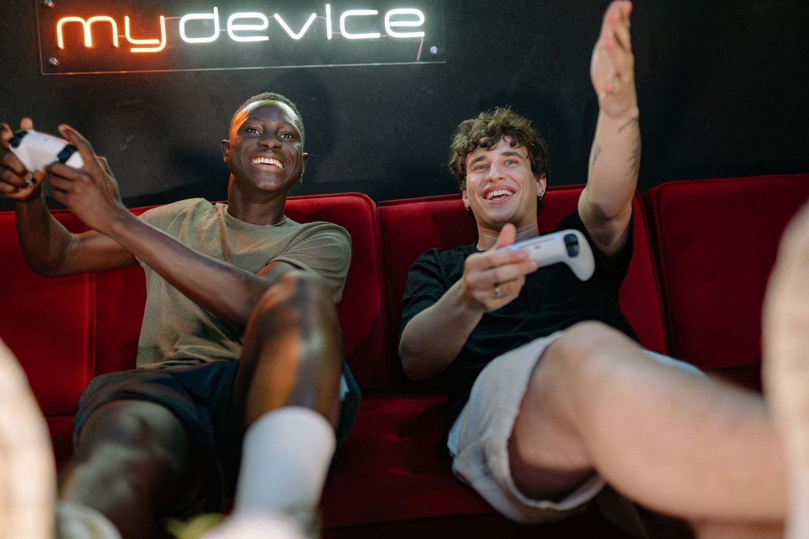 Two young men laughing on a red velvet sofa holding white game controllers, a neon sign glowing on the dark wall behind them