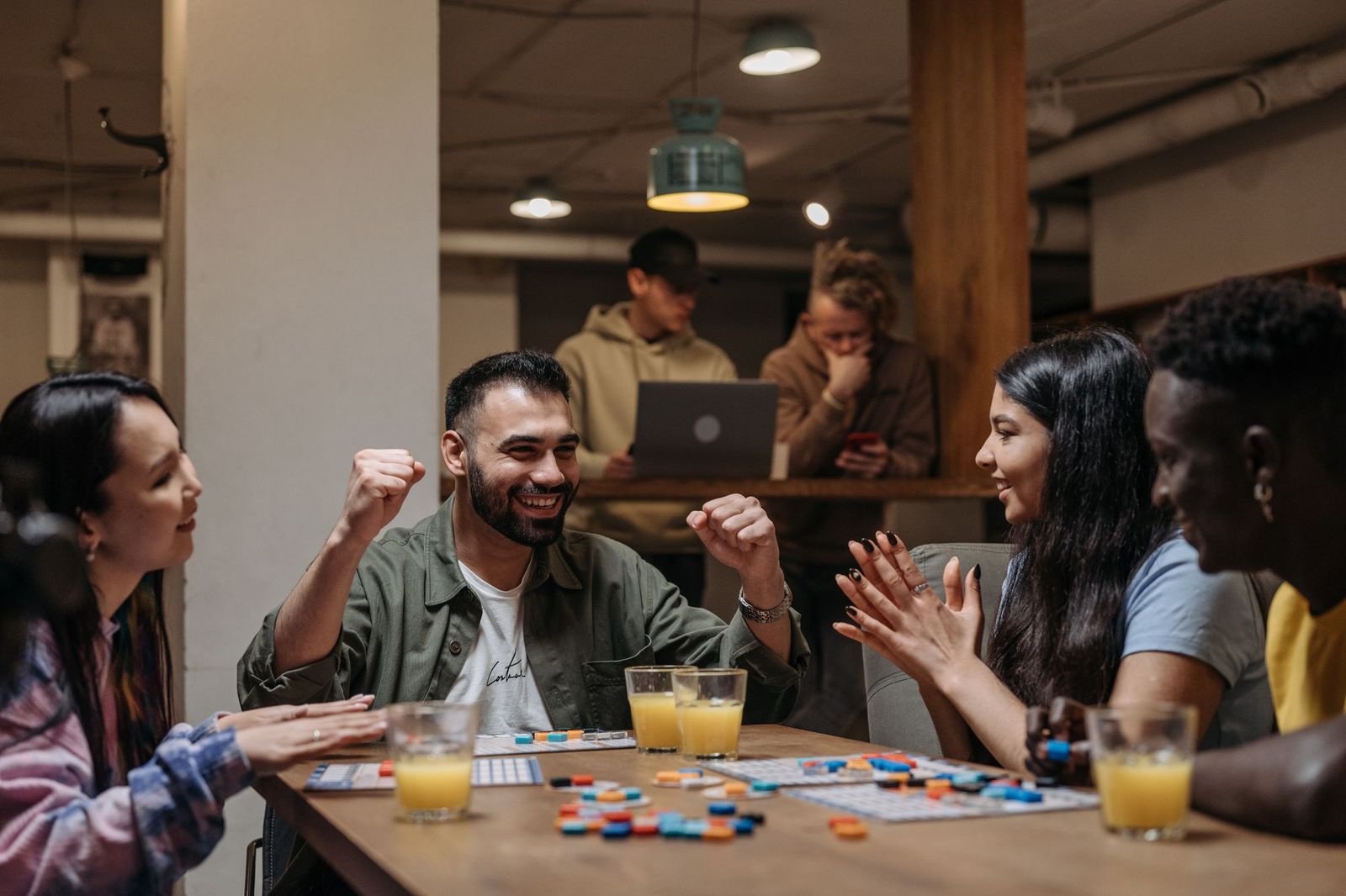 Five friends around a wooden table in a bar playing a colourful tile board game, laughing with glasses of juice beside them