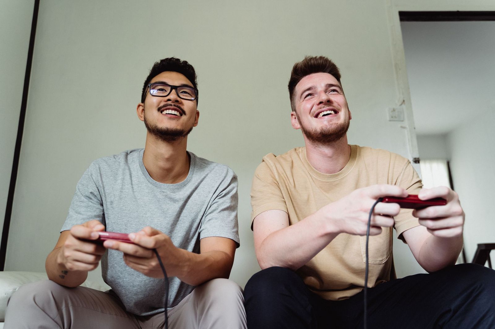 Two friends sitting side by side on a sofa holding game controllers, both laughing and looking up at a screen out of frame