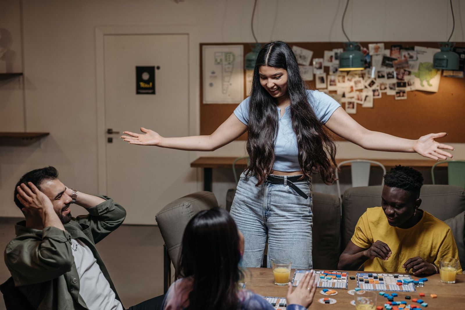 A young woman standing at a table with her arms thrown wide open, smiling, while three seated friends laugh around a half-finished board game