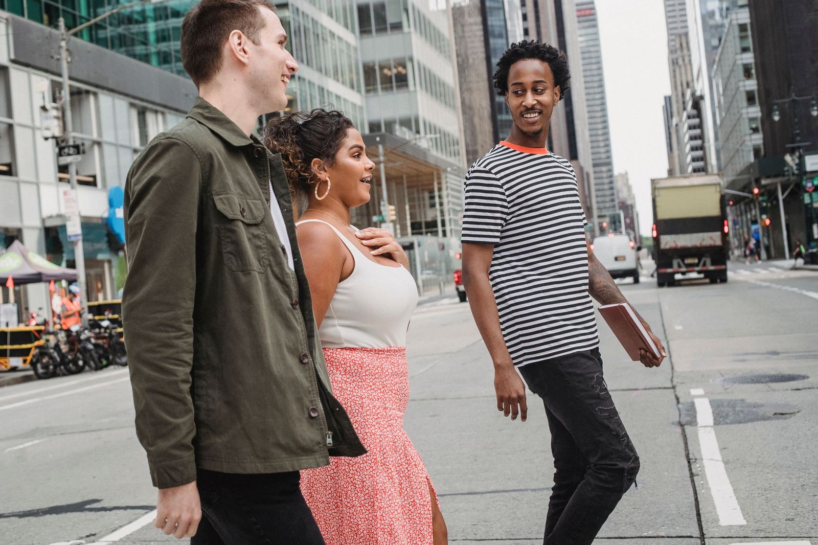 Three friends walking together across a wide city street between tall office buildings, talking and looking at each other