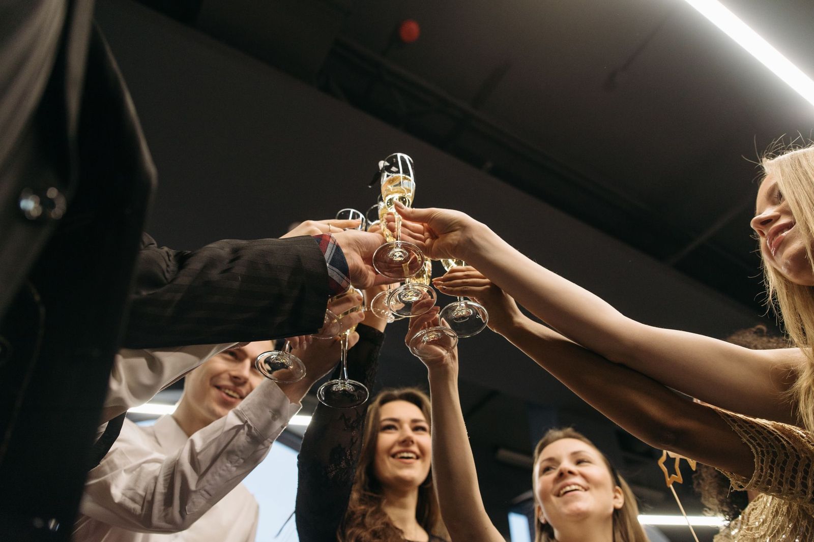 A group of friends photographed from below at night, arms stretched up to clink champagne glasses together under strip lights