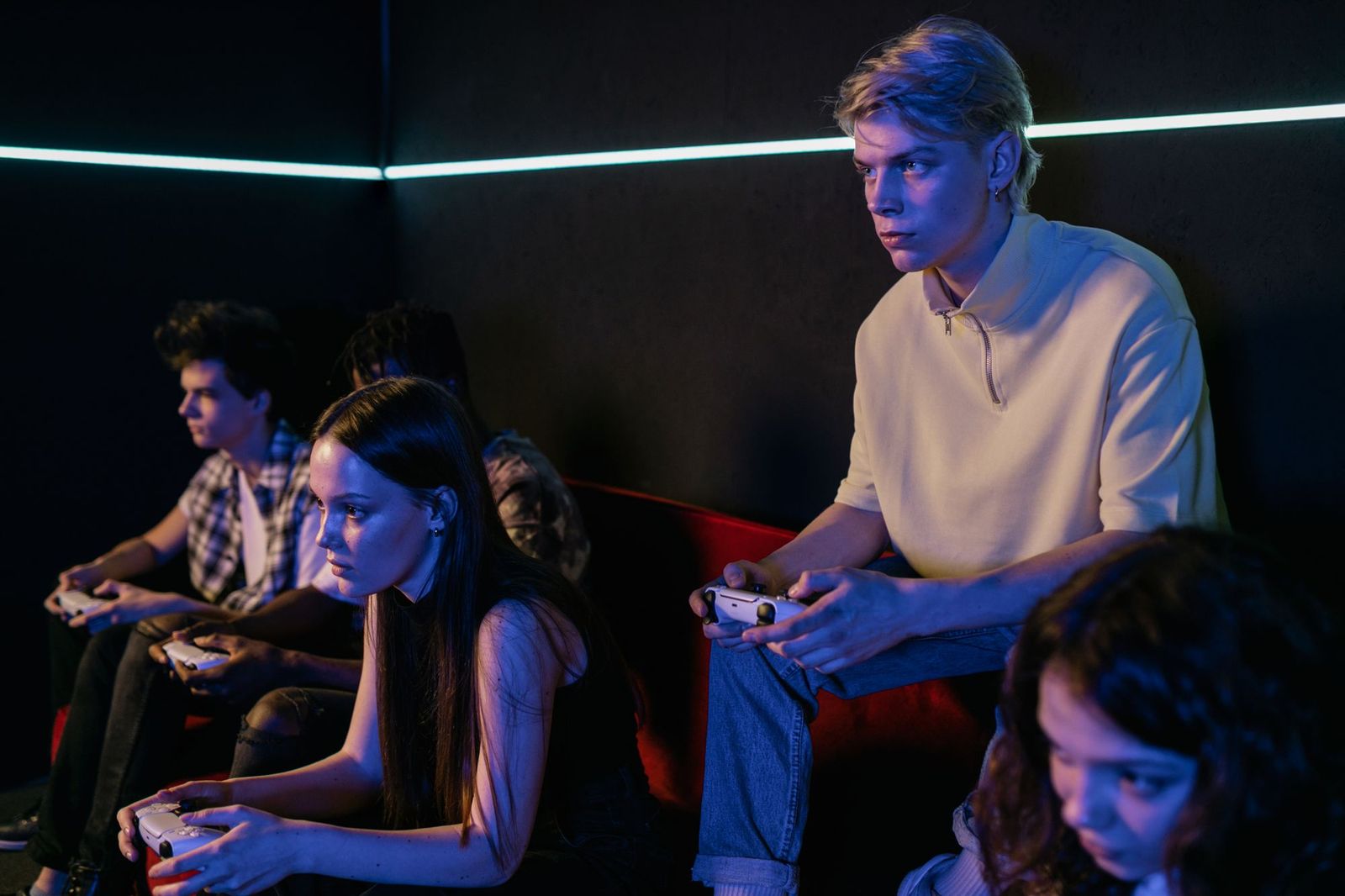 Five young people sitting on a sofa in a dark room lit by neon strips, holding white game controllers and staring at a screen off camera