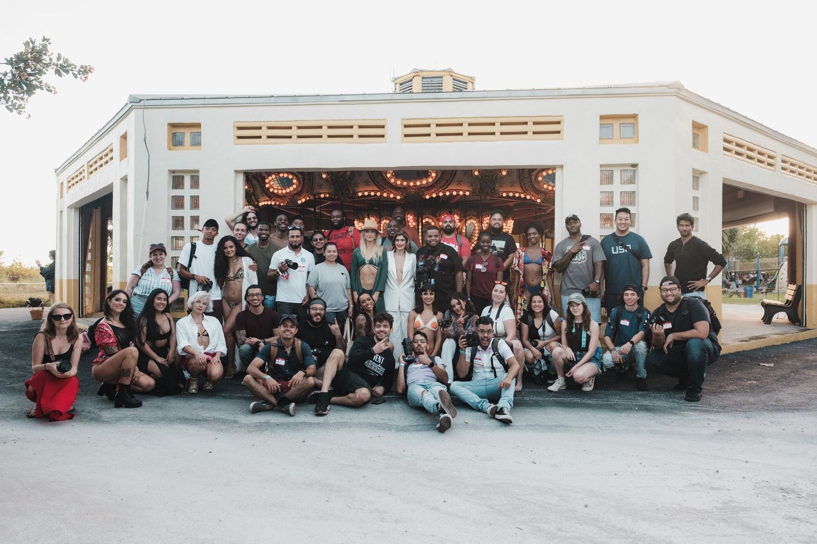 A large group of about forty people posed together in rows outside a pale building with a carousel visible behind them, many wearing name badges and holding cameras