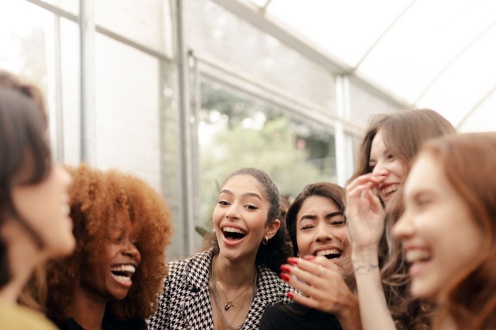 A close group of friends laughing together indoors under a glass roof, heads tilted towards each other