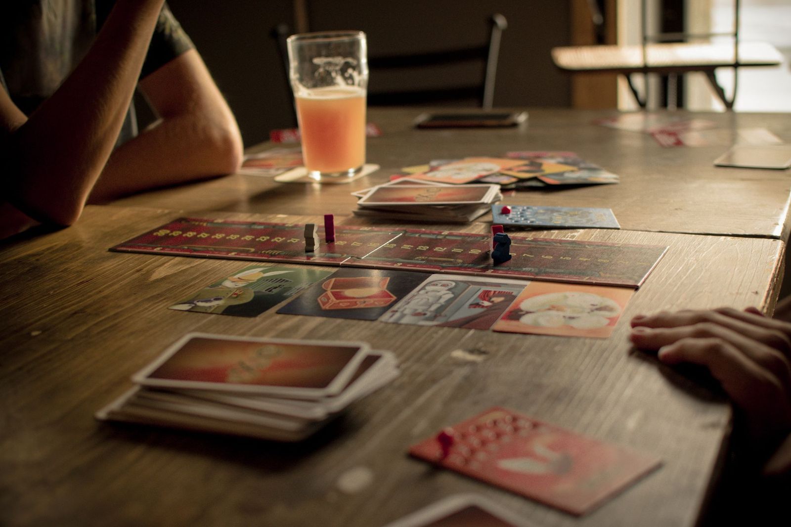 A board game spread across a wooden table in a bar, with cards, small wooden pieces and a half-finished glass of drink beside a player's arms