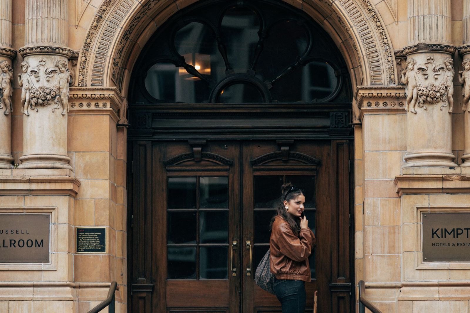 A woman in a brown leather jacket standing at the tall carved stone entrance of a grand old hotel, looking back over her shoulder