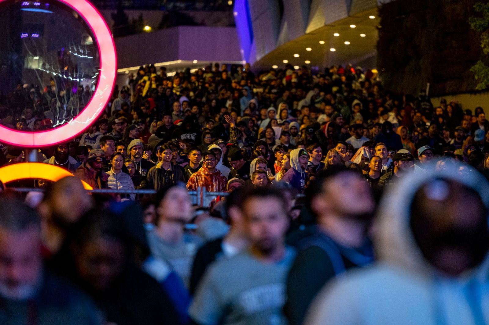 A dense crowd of people packed together outdoors at night, lit by pink and orange neon rings, all facing the same direction