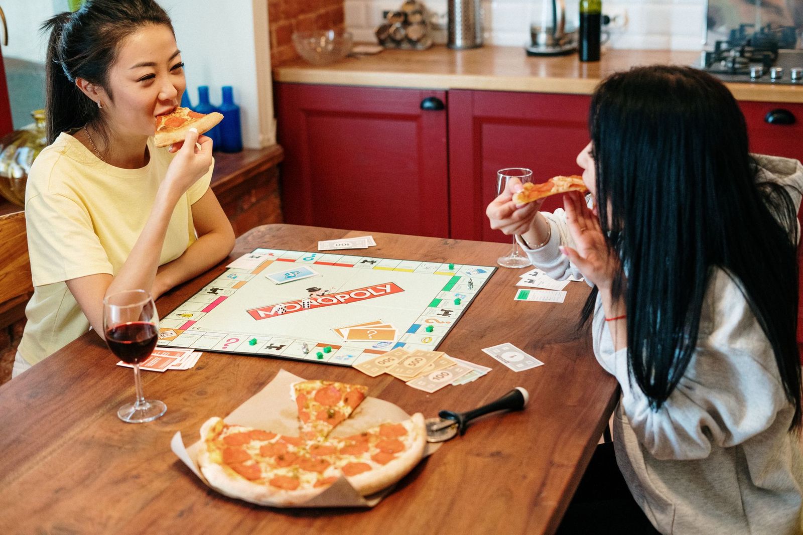 Two friends laughing over slices of pepperoni pizza at a wooden kitchen table with a Monopoly board and play money spread between them