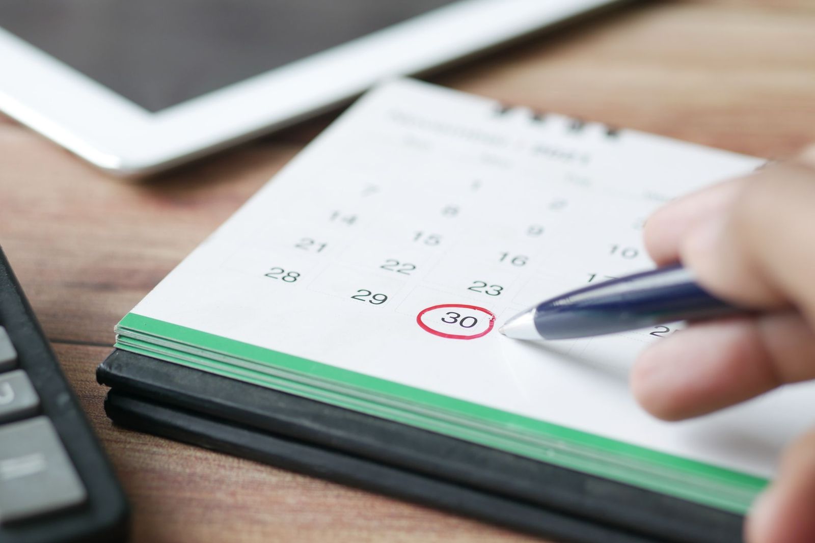 A close-up of a hand circling the 30th on a paper calendar with a pen, a tablet resting nearby
