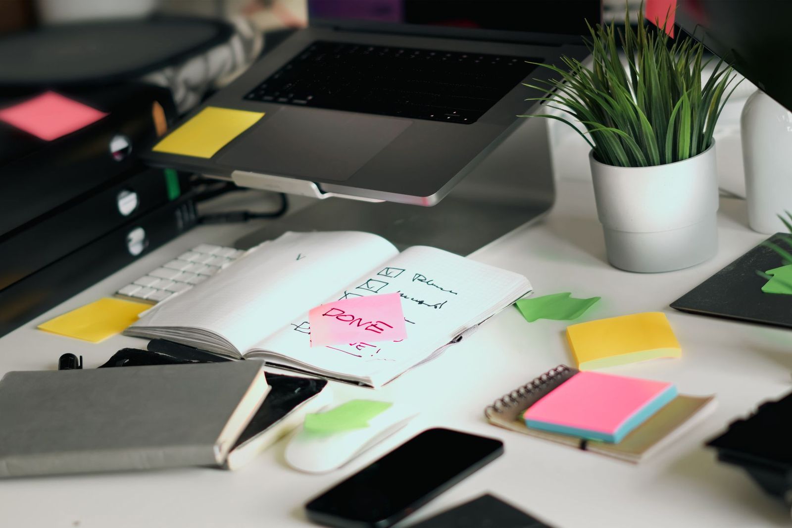 A cluttered desk with a laptop on a stand, colorful sticky notes, a notebook with a checklist and a DONE note, a phone and a small potted plant