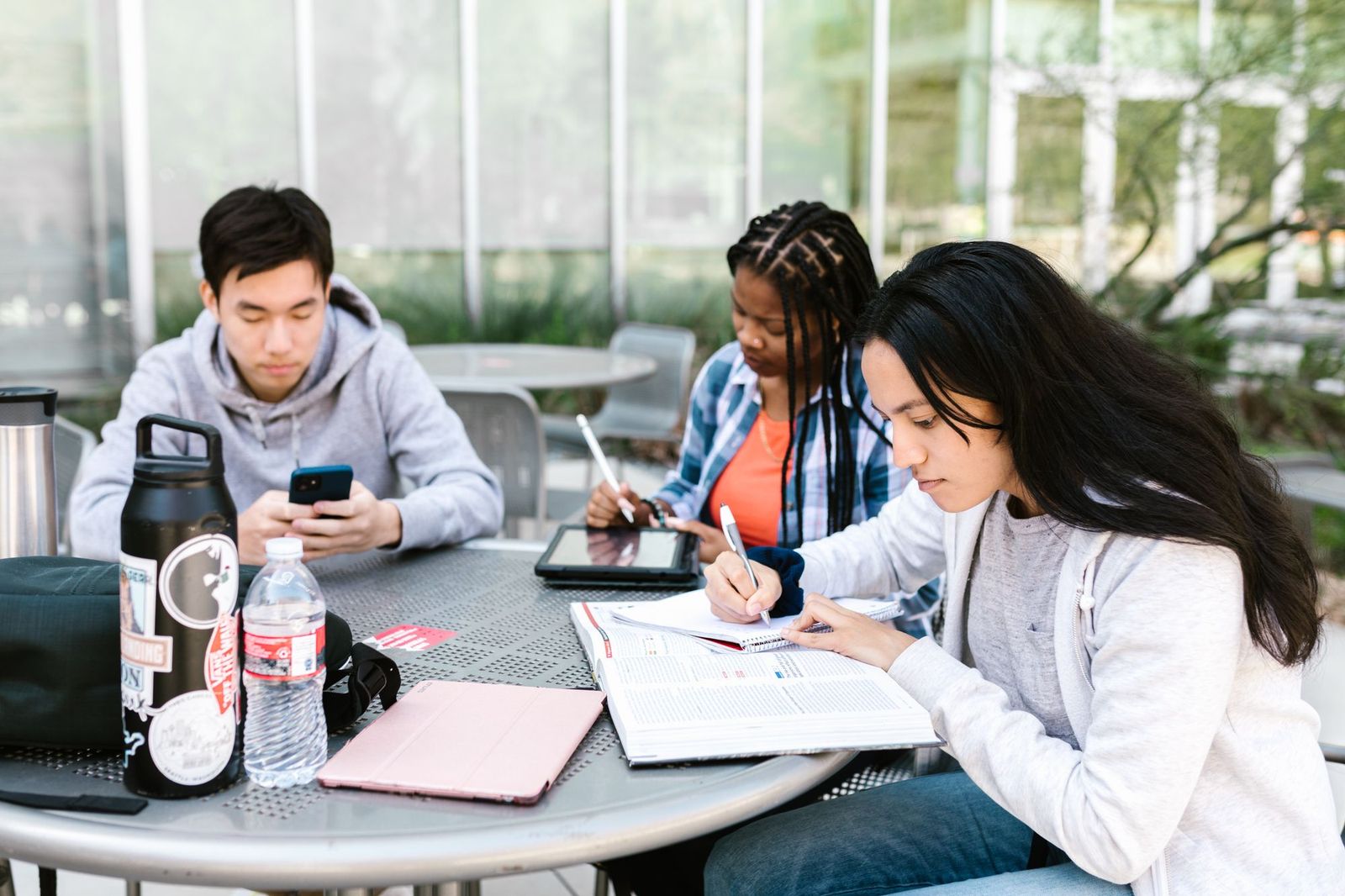 Three friends at an outdoor table, one checking a phone while the other two work through a notebook and a tablet