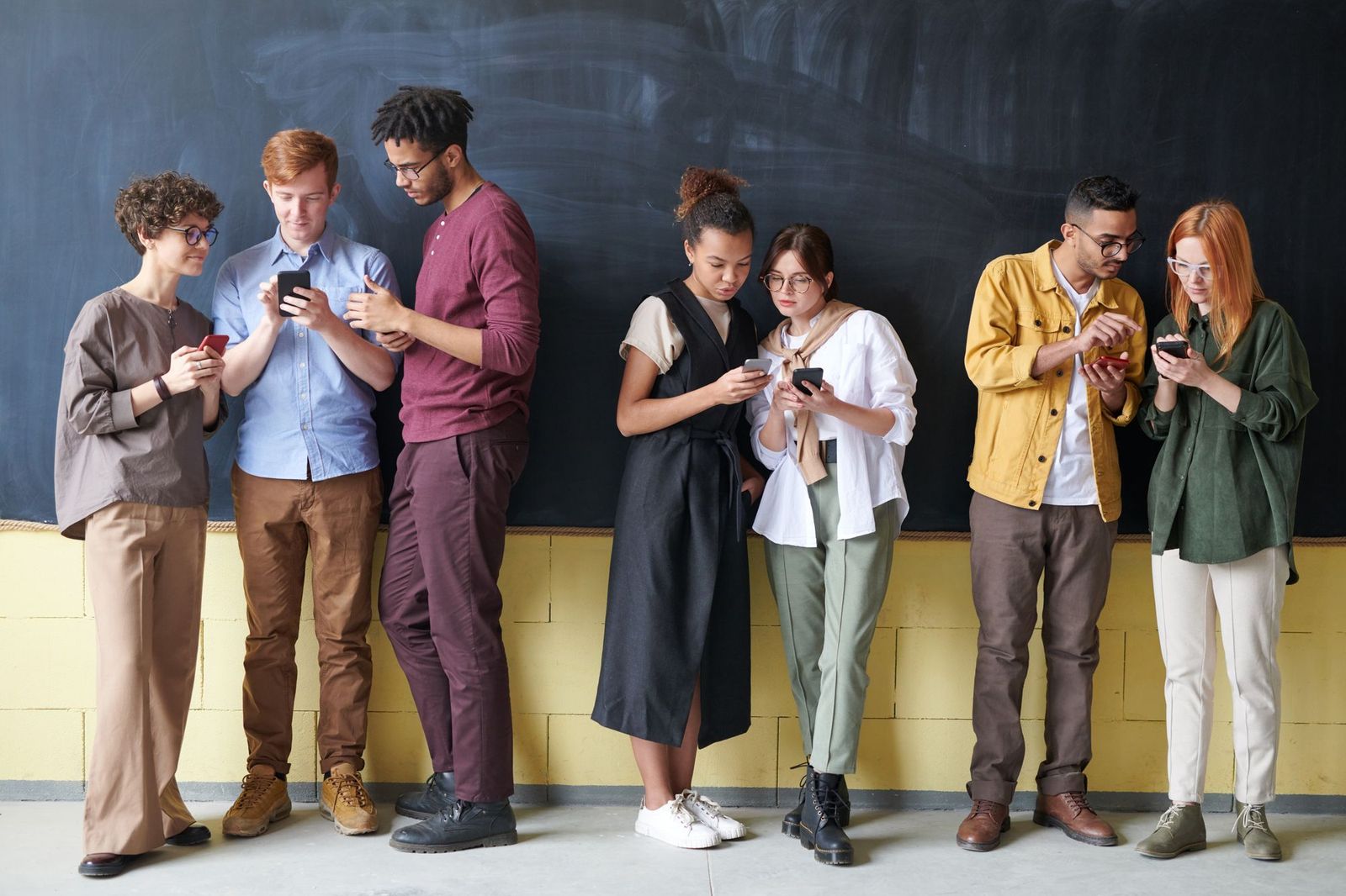 Seven friends in a line against a chalkboard wall, each absorbed in their own phone