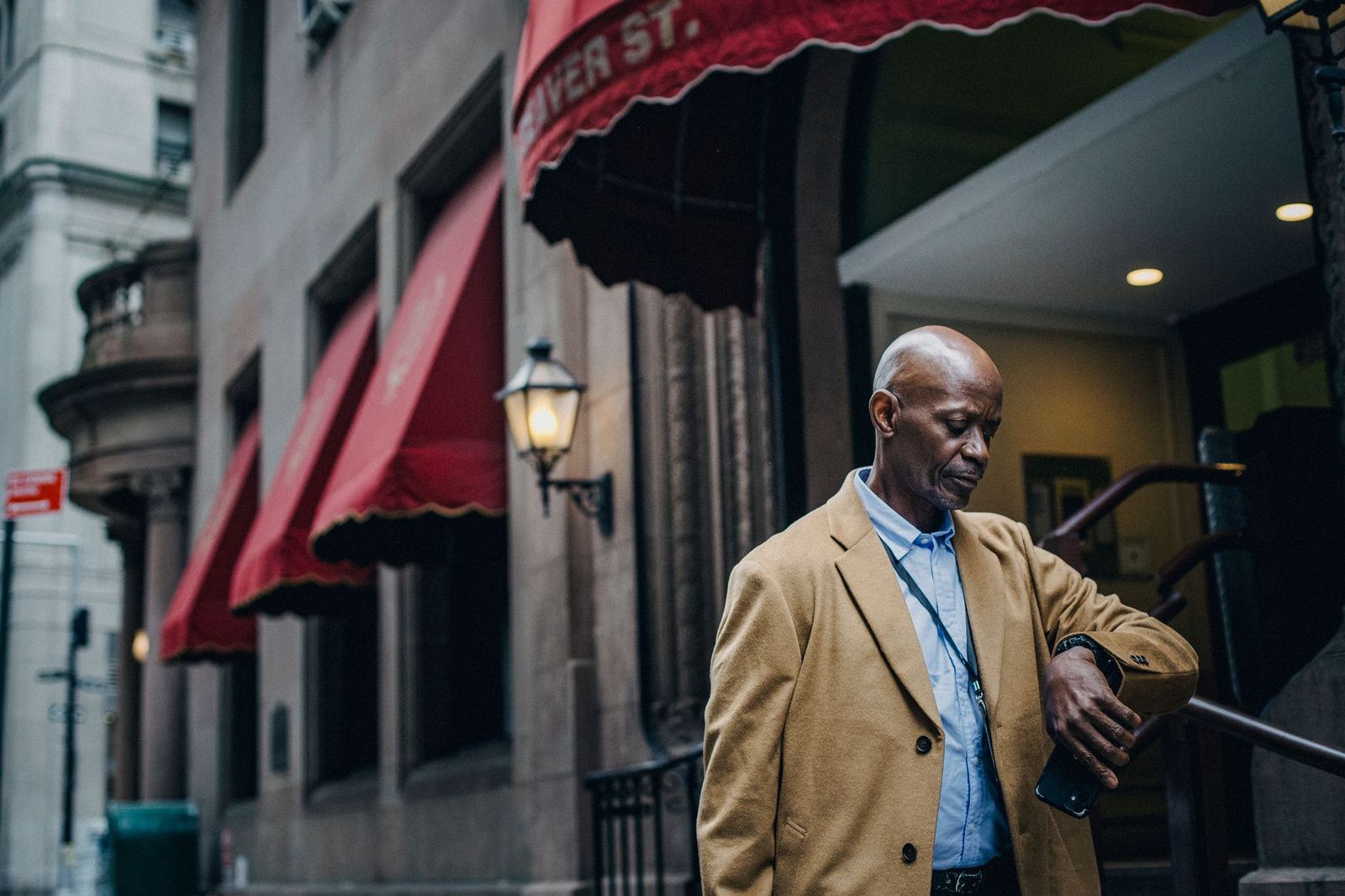 A man in a camel coat pausing outside a building with red awnings on a city street, looking down at his wrist while holding a phone