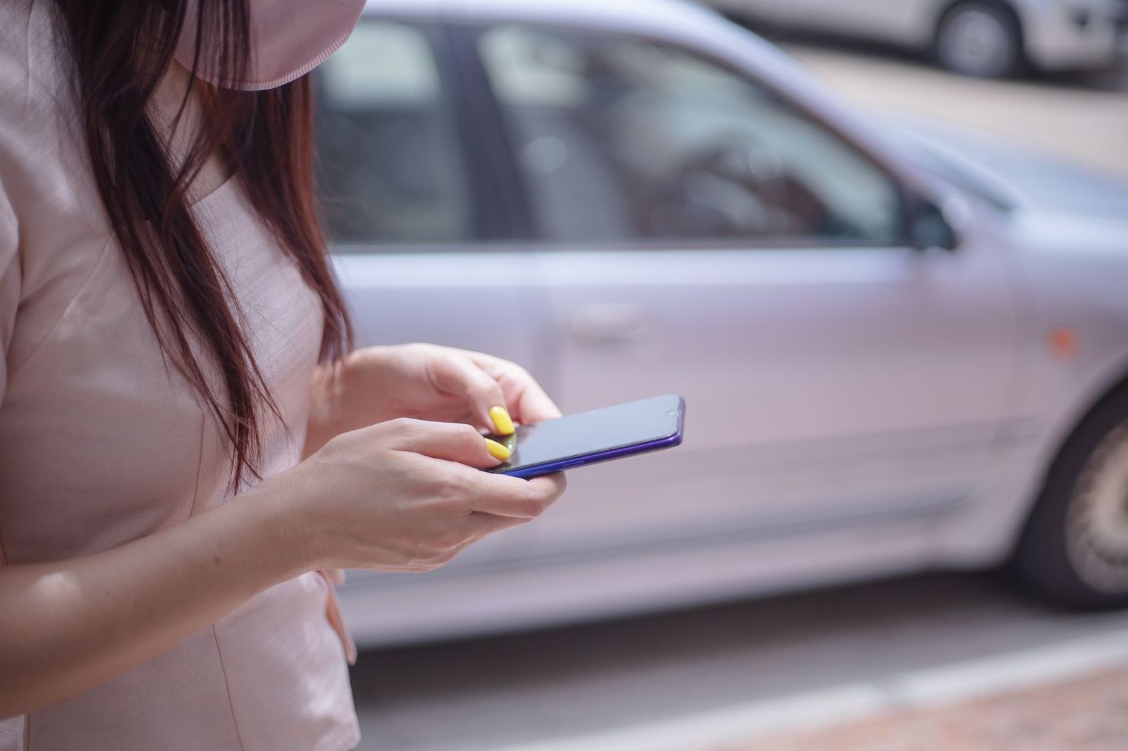 A woman standing on a pavement beside a parked car, holding a phone in both hands and looking down at the screen