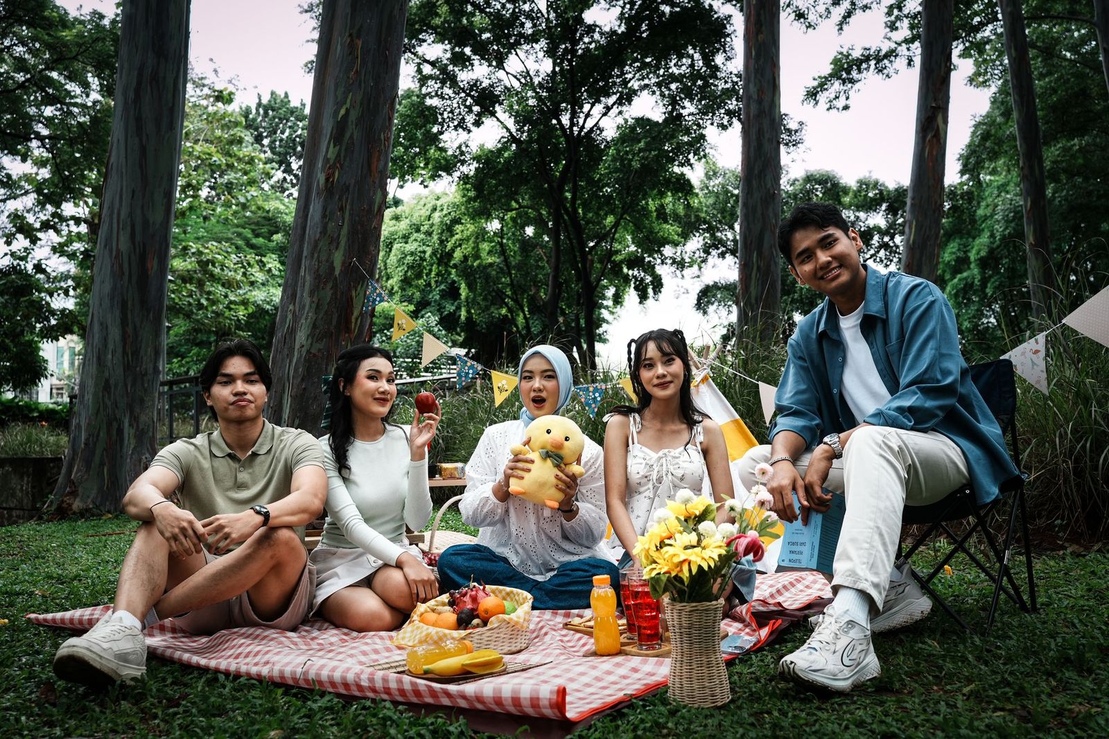 Five friends sitting on a red checked picnic blanket under tall trees in a park, with fruit, flowers, drinks and bunting strung behind them