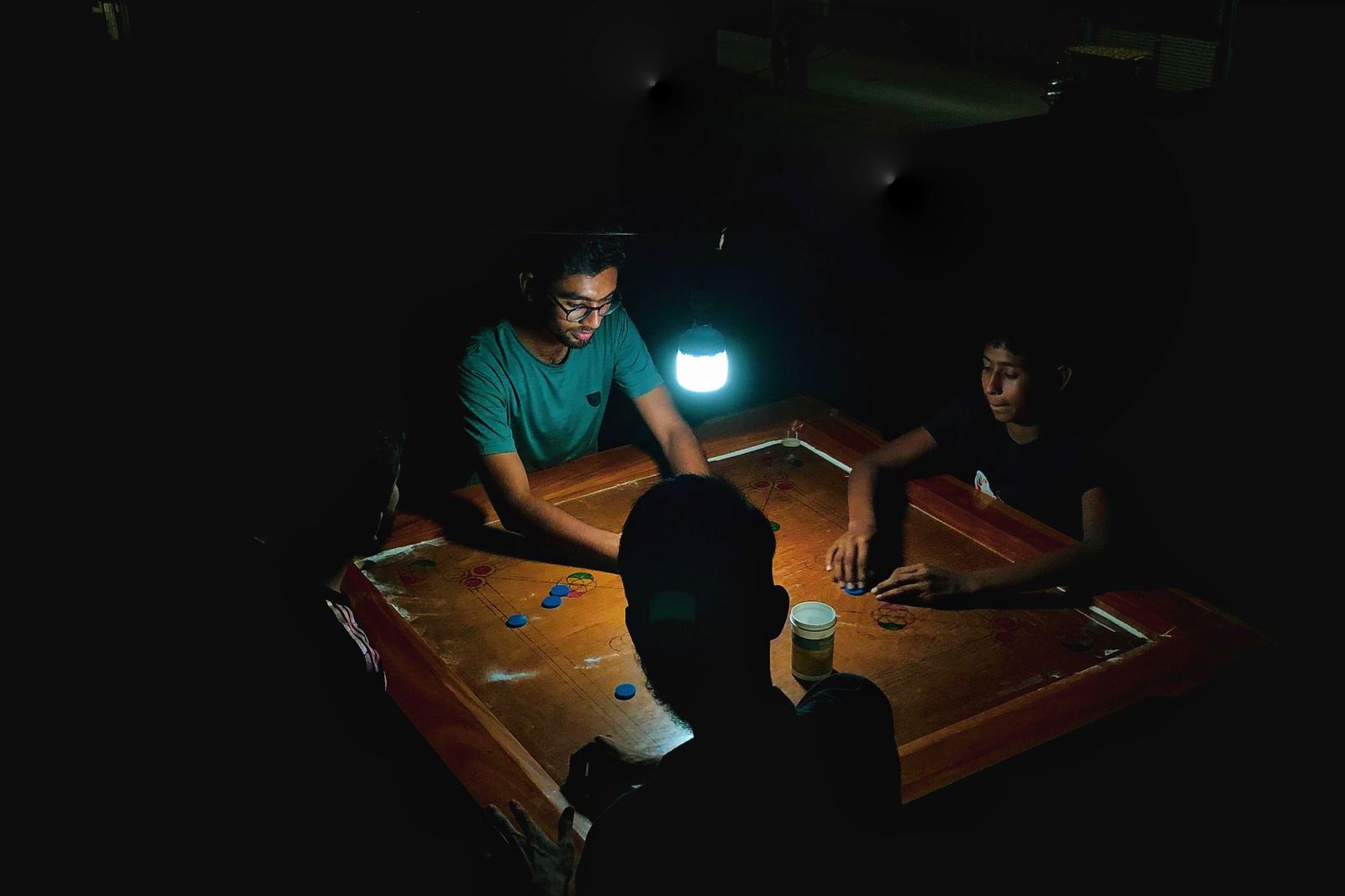 Three people playing a carrom board in a dark room, lit only by a small lantern hanging above the board
