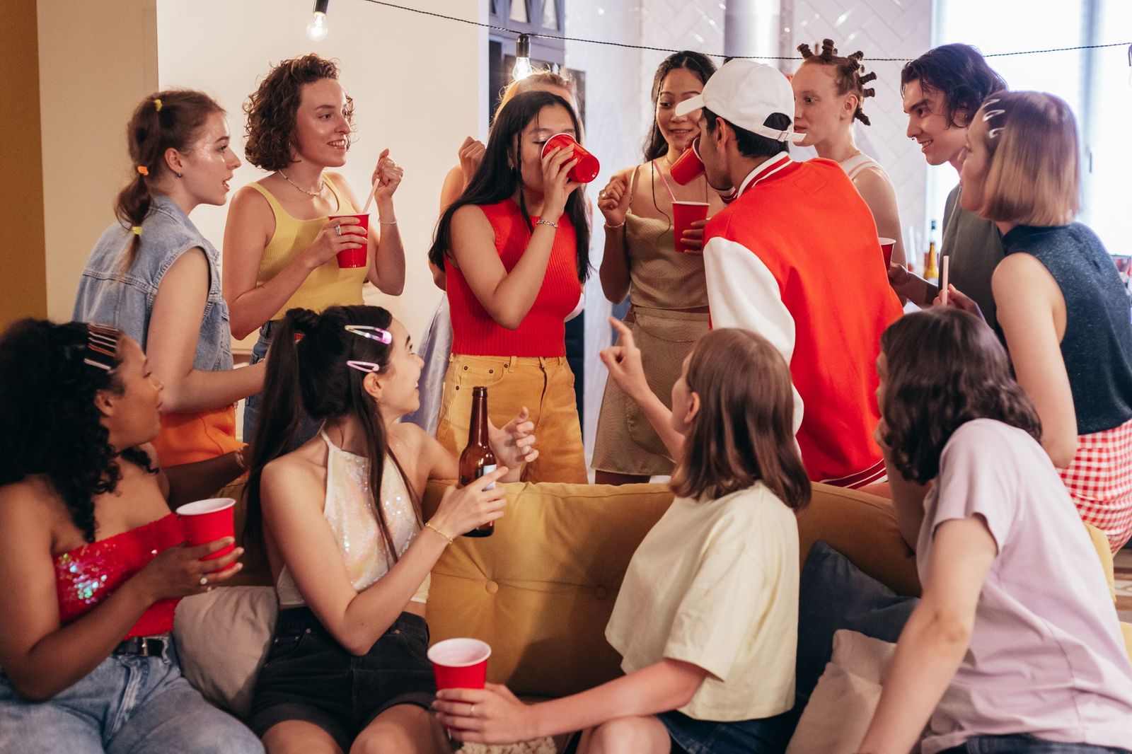 A crowded house party in a bright living room, a dozen young people standing and sitting around a sofa with red cups, talking and laughing