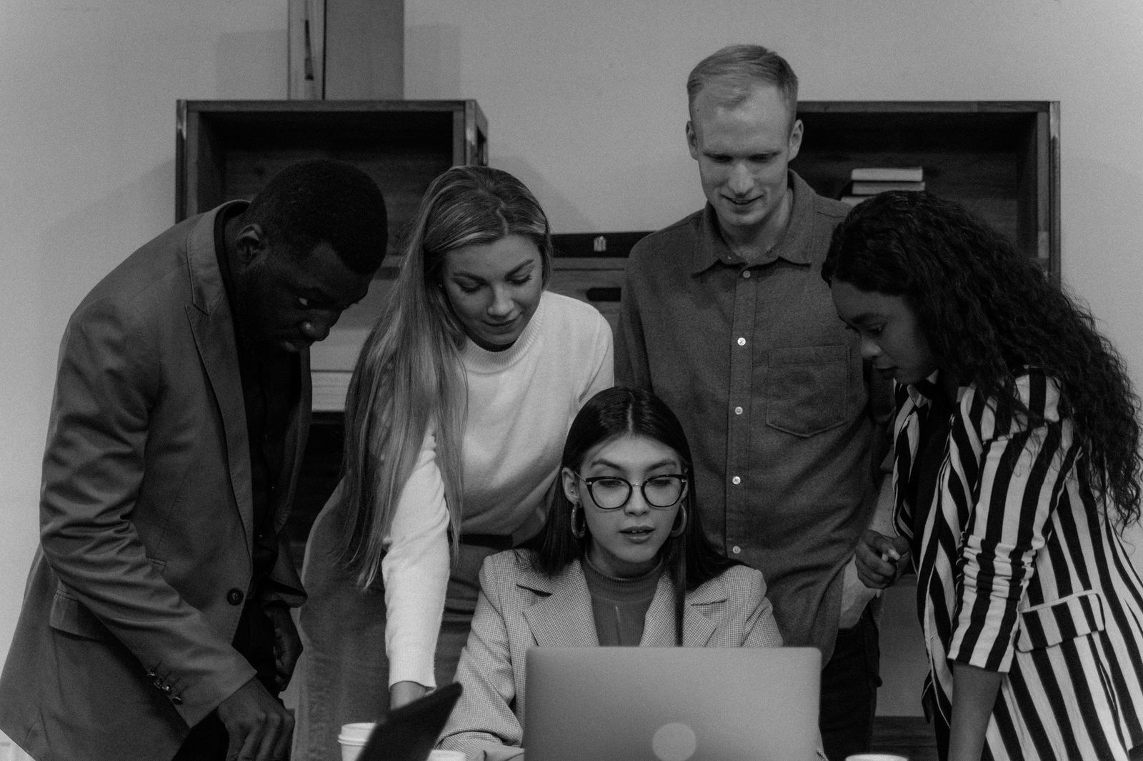 Five colleagues crowded around a single laptop screen, leaning in together, in black and white