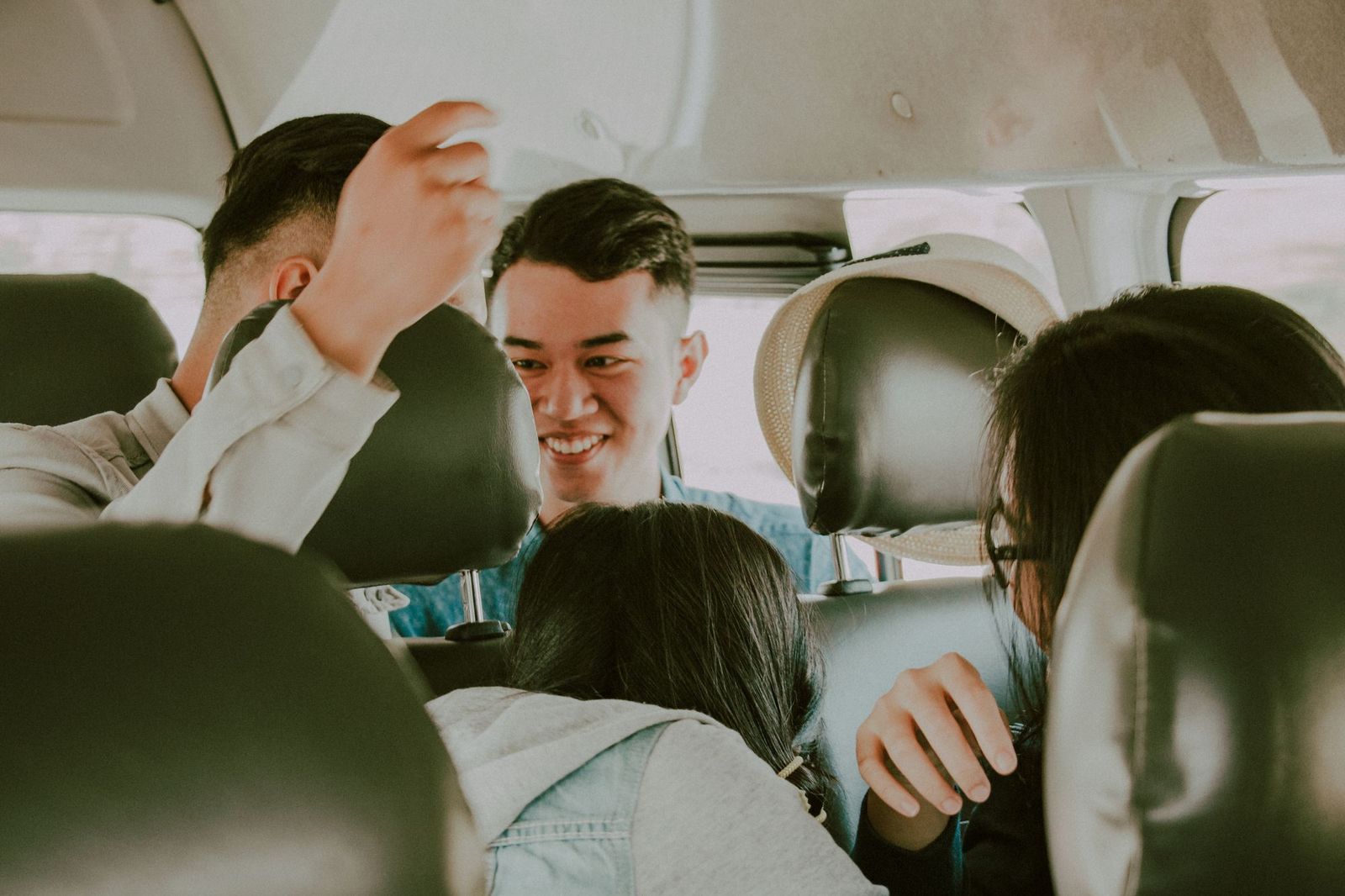 Passengers inside a van seen from the back row between the headrests, one reaching up to the roof and another turning around smiling
