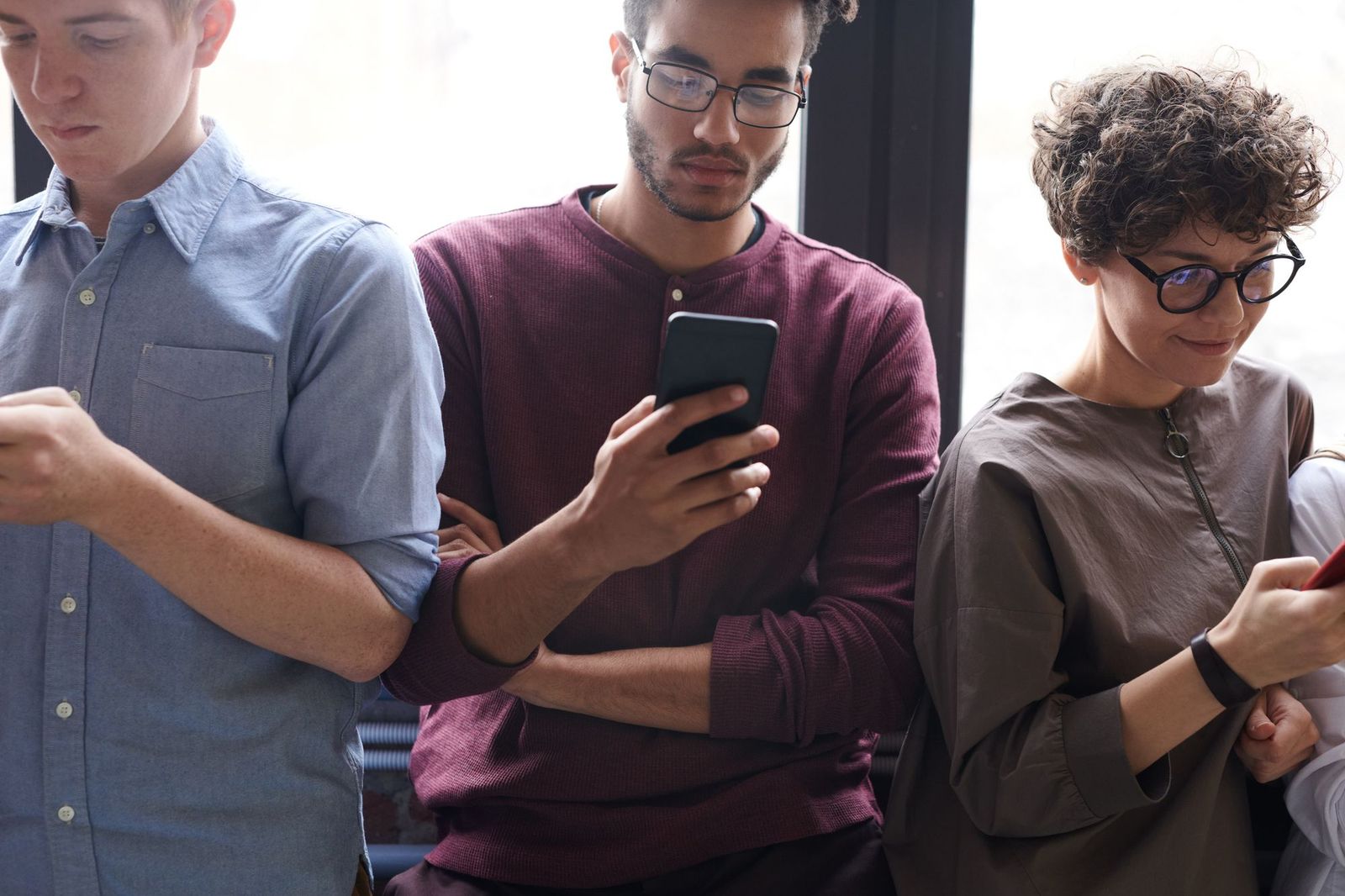Three people standing side by side against a bright window, the one in the middle holding a phone that the others are looking at