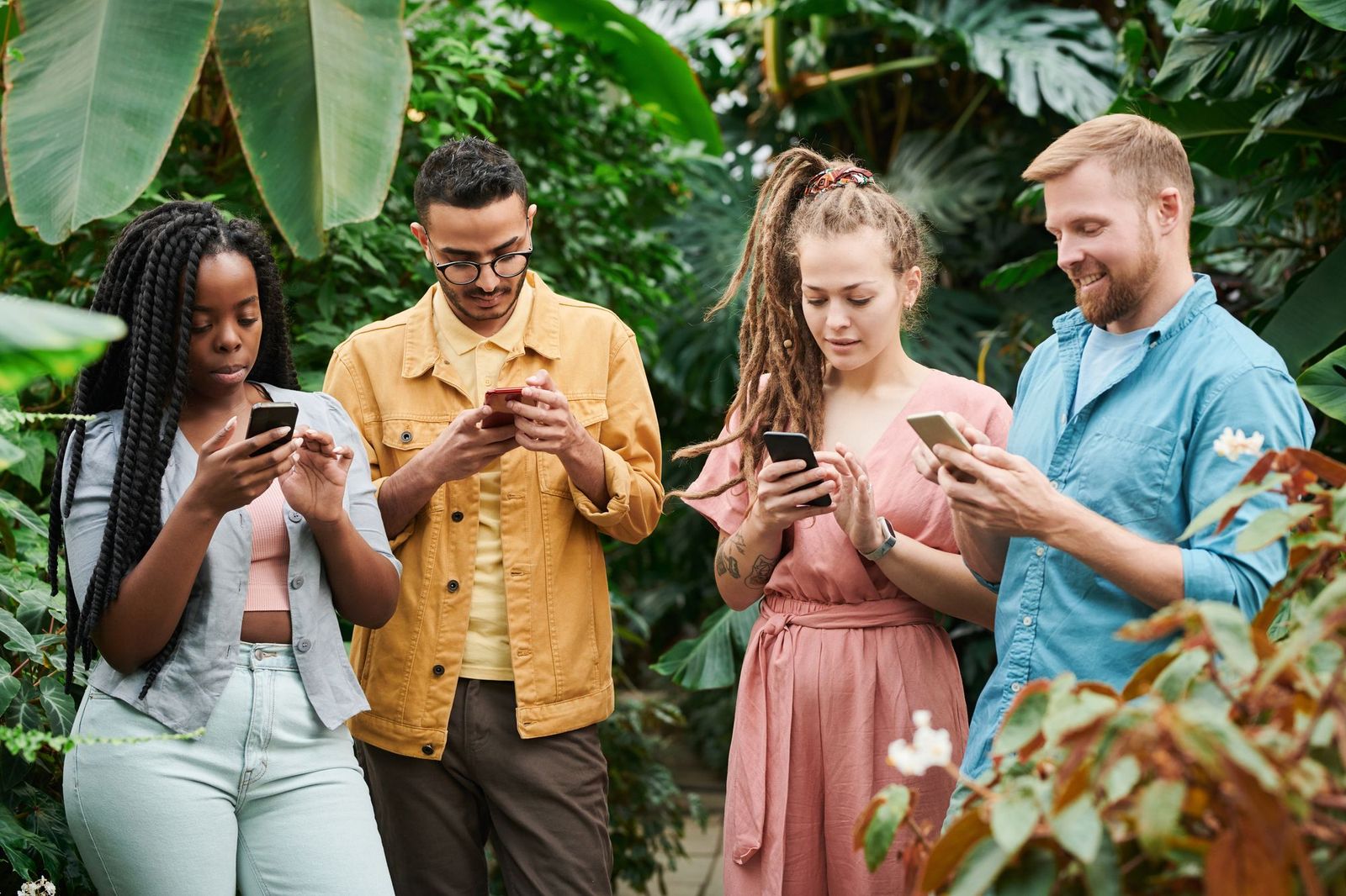Four friends standing side by side outdoors among large green plants, each looking down at their own phone rather than at each other