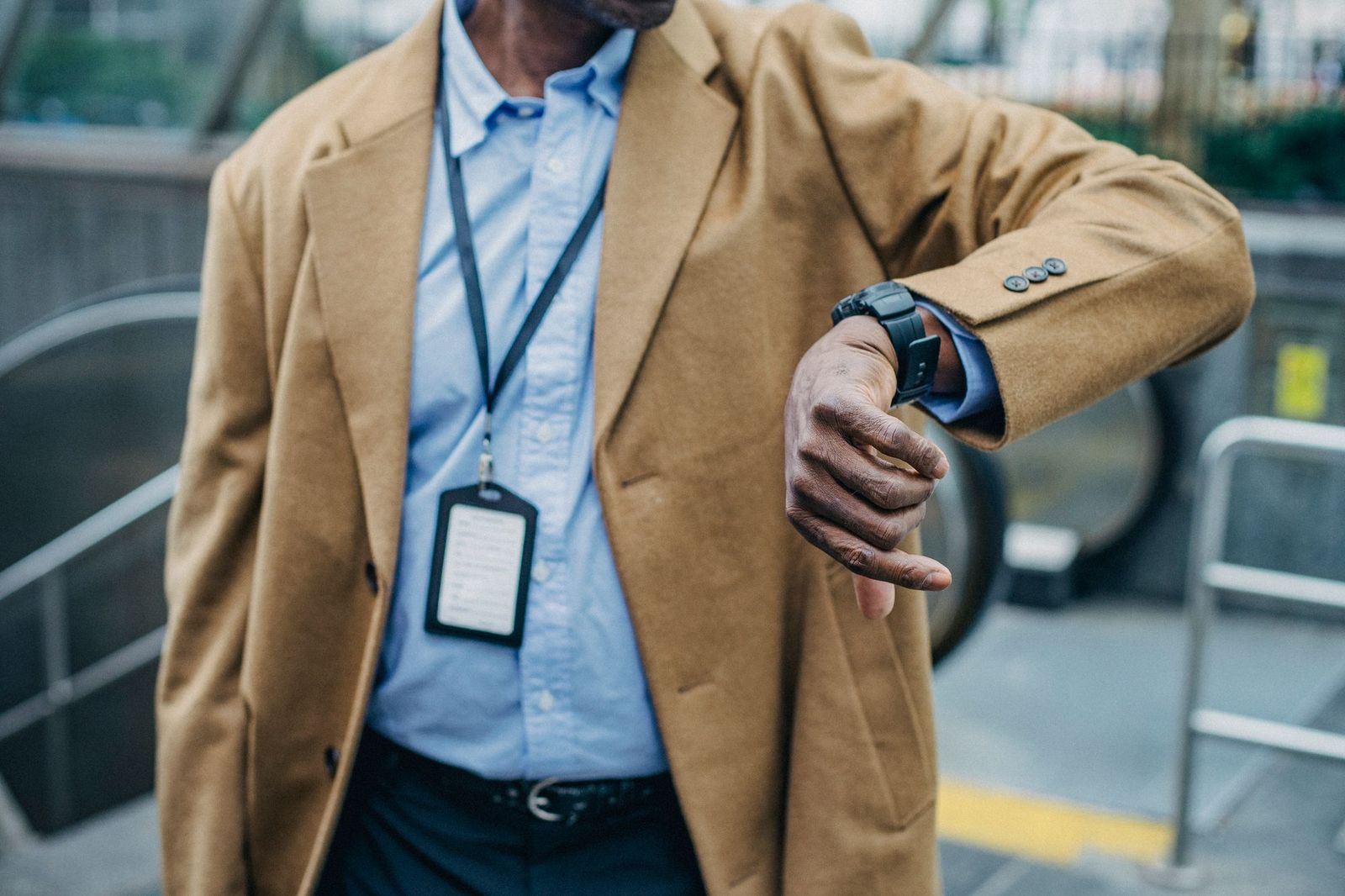 A man in a tan jacket and lanyard checking his wristwatch while standing beside an escalator in a public building