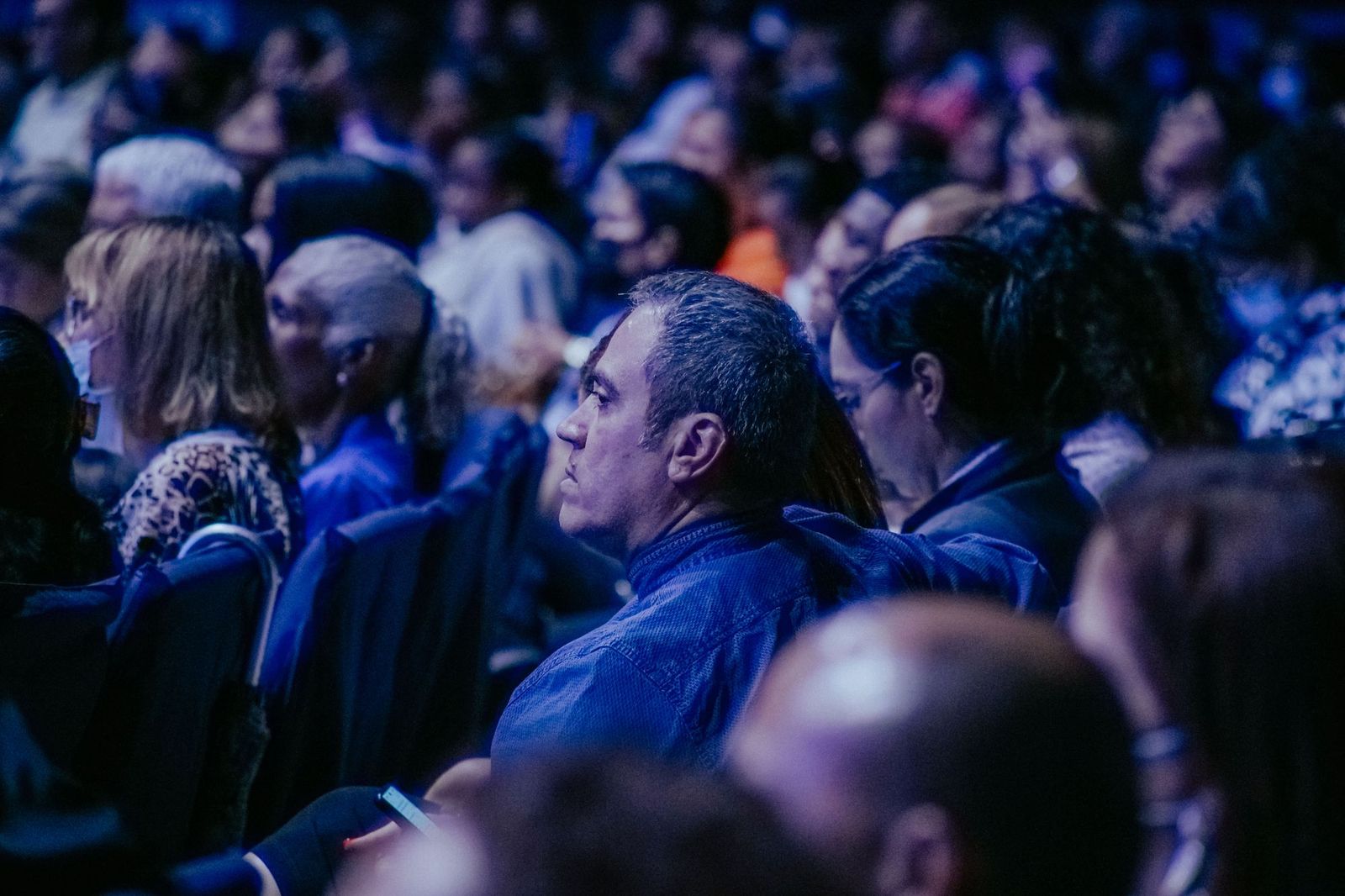 A large audience seated closely together in a dark hall lit in blue, most faces turned towards a stage out of frame