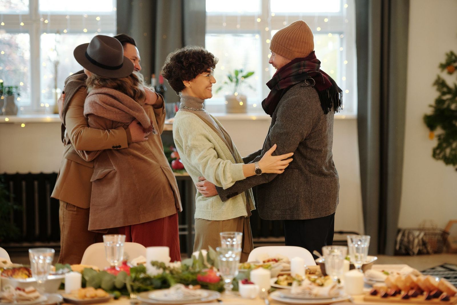 A small group of friends embracing warmly in pairs at an indoor gathering, a decorated dinner table set in the foreground