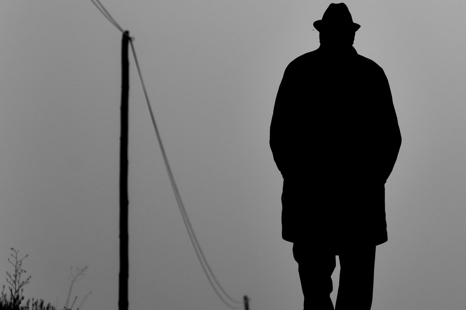 A solitary figure in a coat and hat photographed in silhouette against a grey sky, walking alone past a utility pole and wires