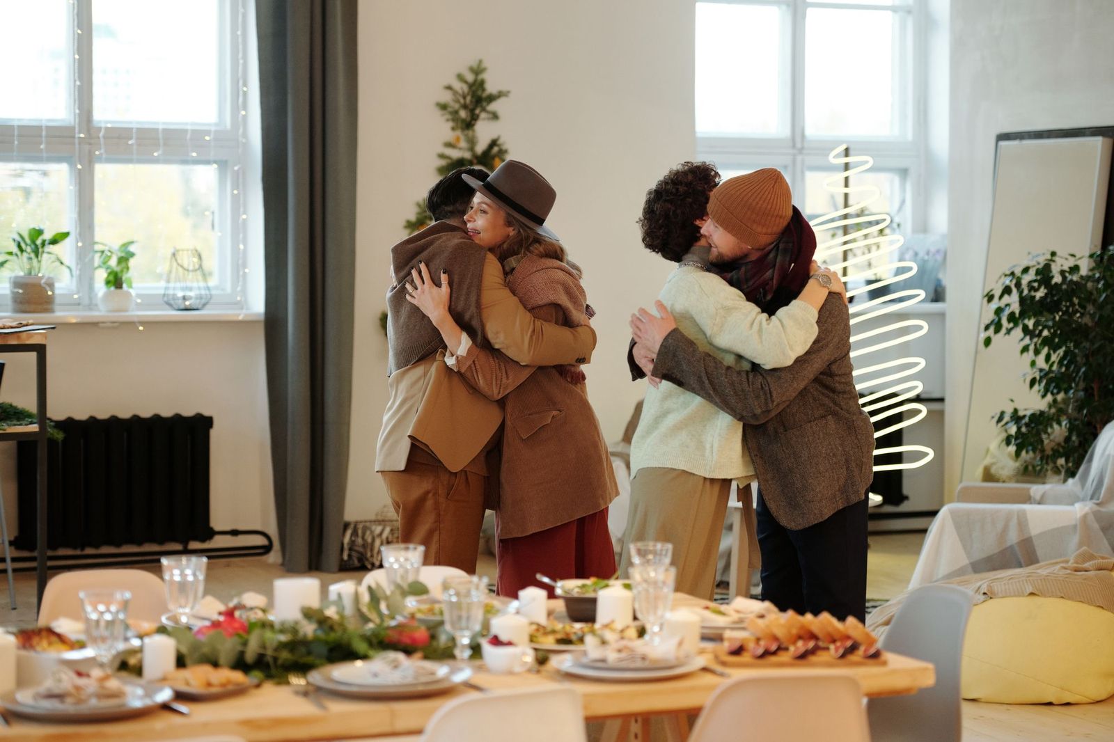 Two pairs of friends embracing in greeting inside a bright apartment, a long table set for a holiday meal in the foreground